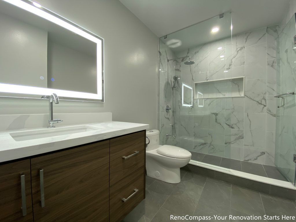 This Oakville bathroom renovation showcases a modern walk-in glass shower with marble-look wall tiles, built-in shower niche, and sleek frameless enclosure for a clean contemporary design. The space also features a custom wood vanity with quartz countertop, LED backlit mirror, and upgraded fixtures that enhance both functionality and visual appeal. Completed by a professional Toronto renovation company, this bathroom upgrade focuses on maximizing brightness, storage, and comfort while delivering a stylish and durable modern bathroom space.