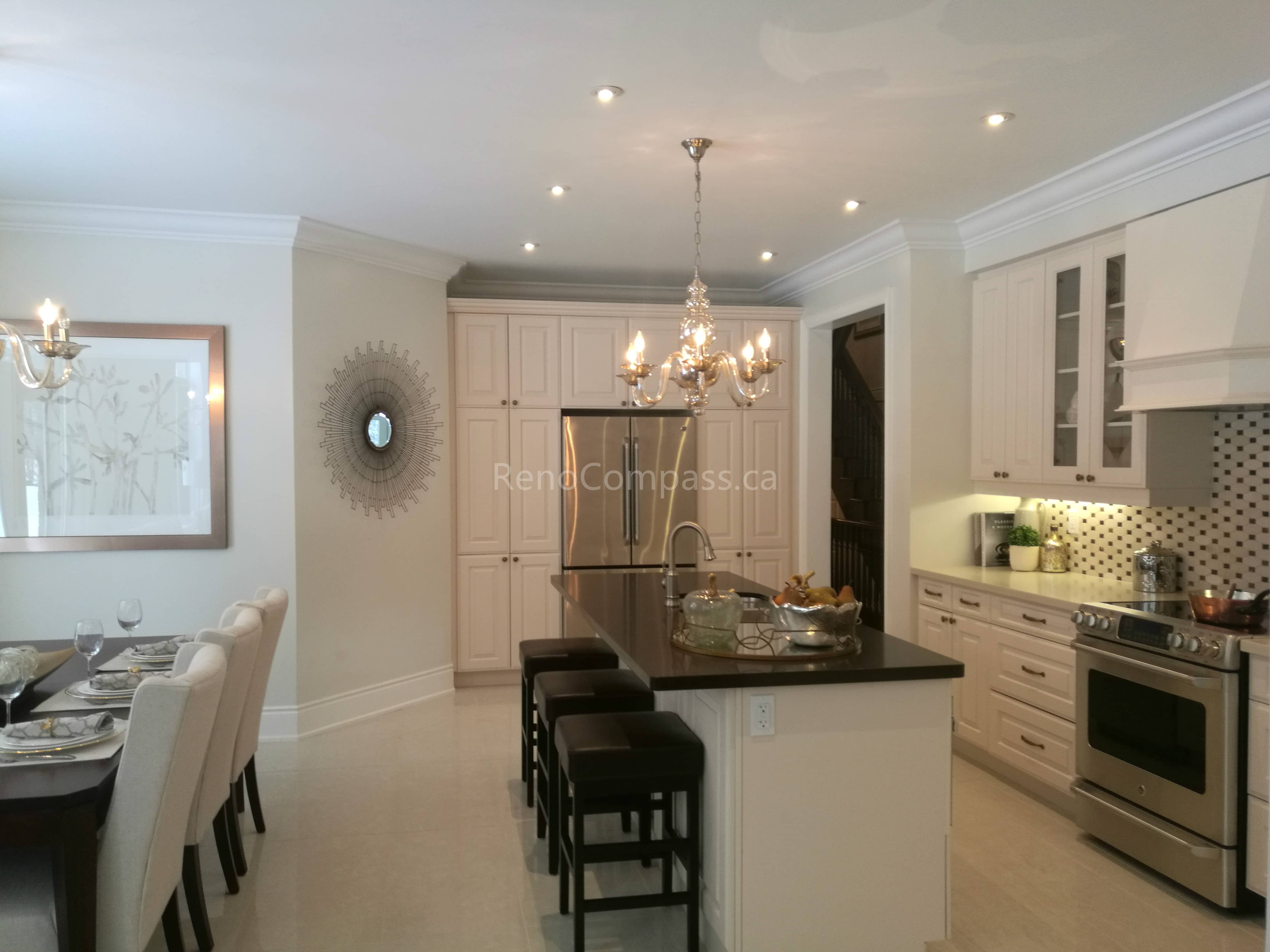 This image highlights an open-concept kitchen and dining area completed as part of a Toronto interior renovation project. The space features custom white cabinetry, a large central kitchen island with seating, and elegant recessed ceiling lighting combined with a classic chandelier. Dark stone countertops contrast beautifully with the light cabinetry, enhancing both functionality and visual balance. The seamless connection between the kitchen and dining area creates an inviting layout ideal for everyday living and entertaining, showcasing refined craftsmanship and timeless interior renovation design for Toronto homes.