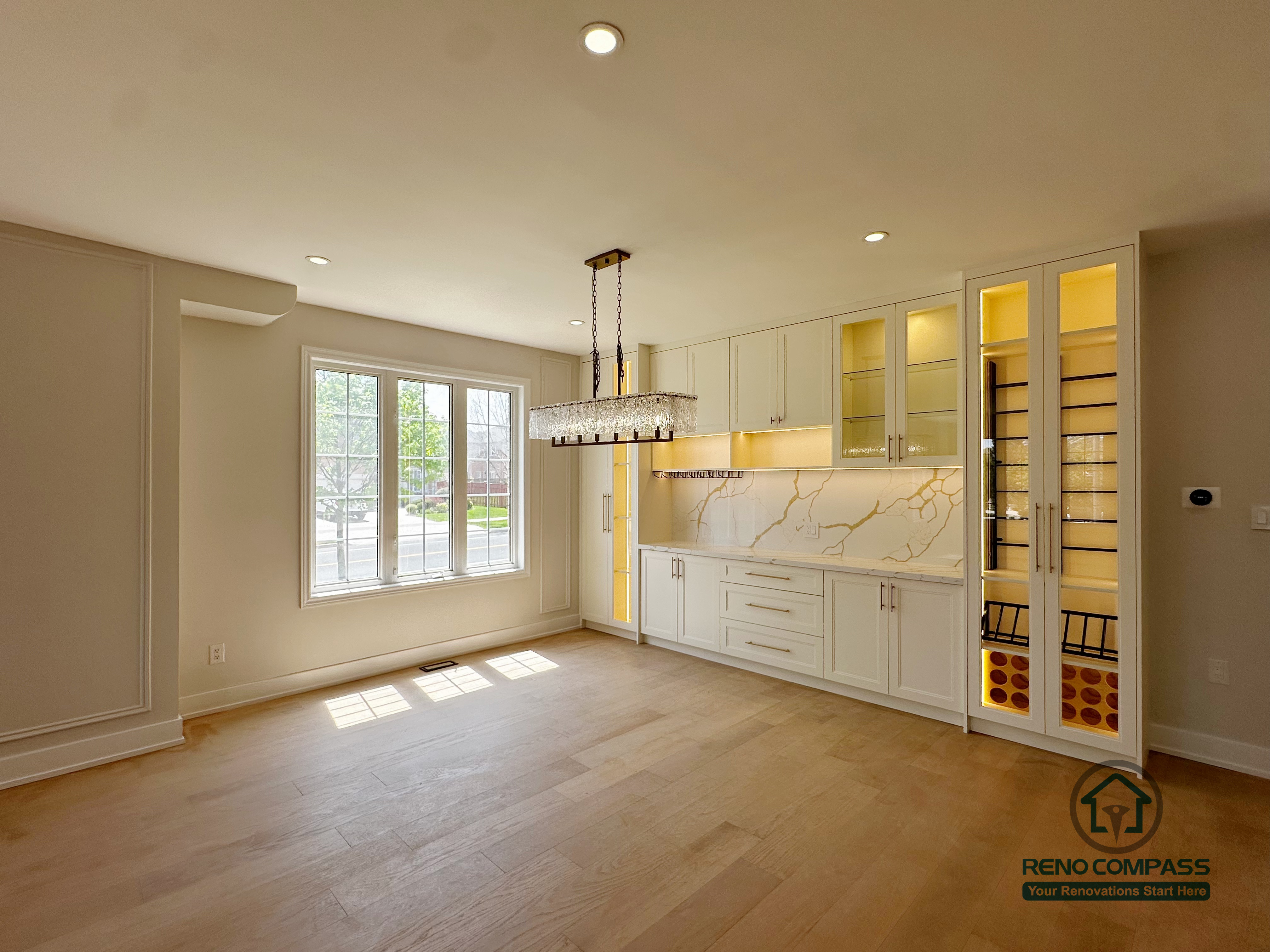 This image features the dining area from a Markham interior renovation, where custom cabinetry and spatial balance were carefully integrated into the overall design. A full-height built-in cabinet wall provides both storage and display functions, enhanced by warm interior lighting that adds depth and visual interest without overpowering the space.

The marble-look backsplash introduces subtle texture and elegance, while the clean white cabinetry keeps the room bright and refined. A statement chandelier anchors the dining area, clearly defining the function of the space while maintaining openness and visual continuity with the surrounding living areas.

Natural light from the large window complements the neutral palette and light wood flooring, creating a comfortable and inviting atmosphere for everyday dining and gatherings. This renovation reflects Reno Compass’s approach to interior design—combining practical storage solutions with cohesive aesthetics for modern Markham homes.