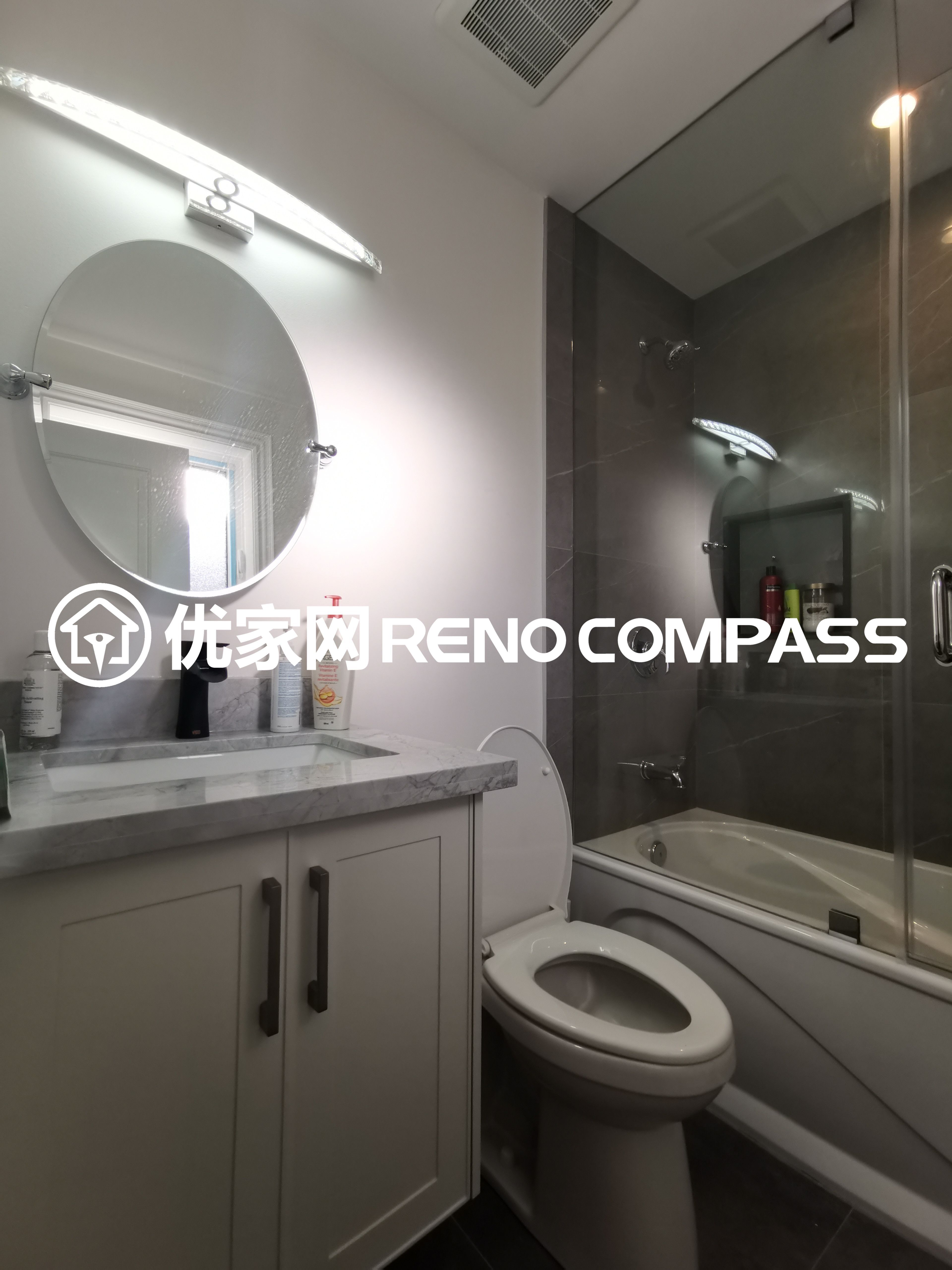 This image highlights a fully renovated bathroom completed as part of a North York interior renovation project. The space features a custom vanity with a marble countertop, paired with a round mirror and integrated vanity lighting that enhances brightness and daily usability. A glass-enclosed bathtub and shower combination maximizes space efficiency while maintaining a clean, modern aesthetic. Neutral finishes and carefully selected fixtures reflect quality craftsmanship and thoughtful bathroom design, delivering a practical and elegant solution for contemporary Toronto homes.