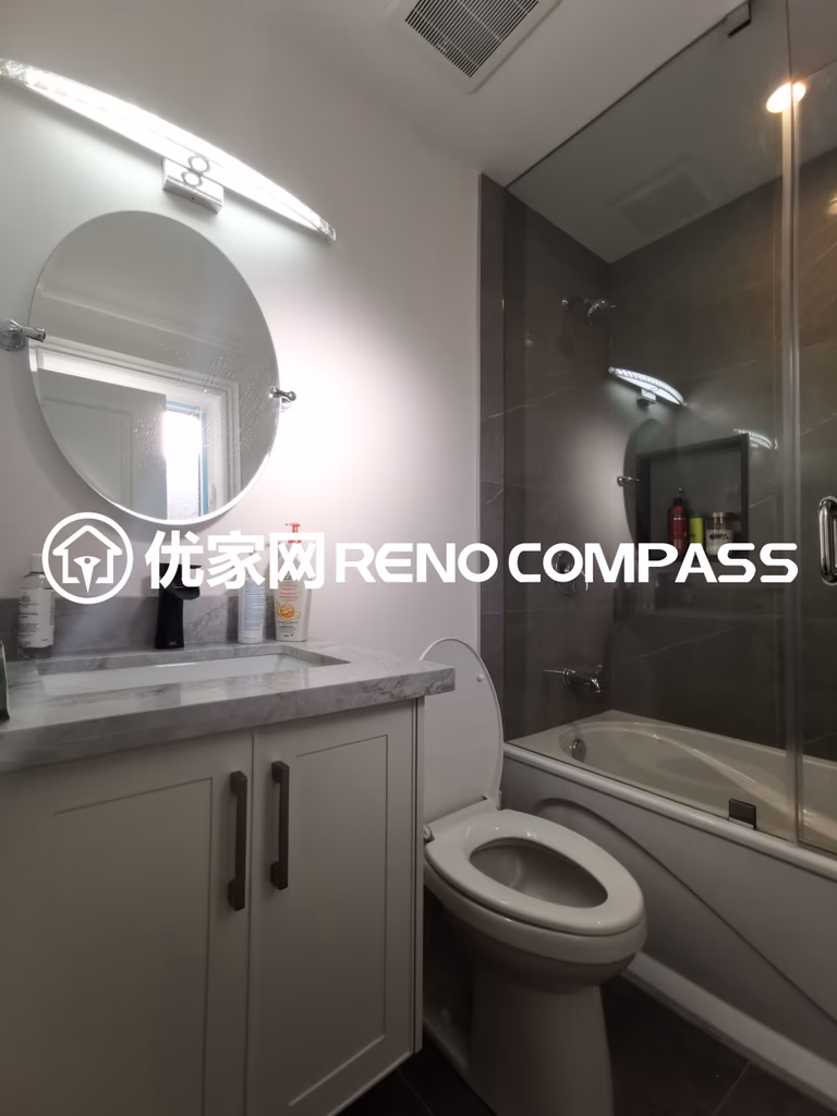 This image highlights a fully renovated bathroom completed as part of a North York interior renovation project. The space features a custom vanity with a marble countertop, paired with a round mirror and integrated vanity lighting that enhances brightness and daily usability. A glass-enclosed bathtub and shower combination maximizes space efficiency while maintaining a clean, modern aesthetic. Neutral finishes and carefully selected fixtures reflect quality craftsmanship and thoughtful bathroom design, delivering a practical and elegant solution for contemporary Toronto homes.