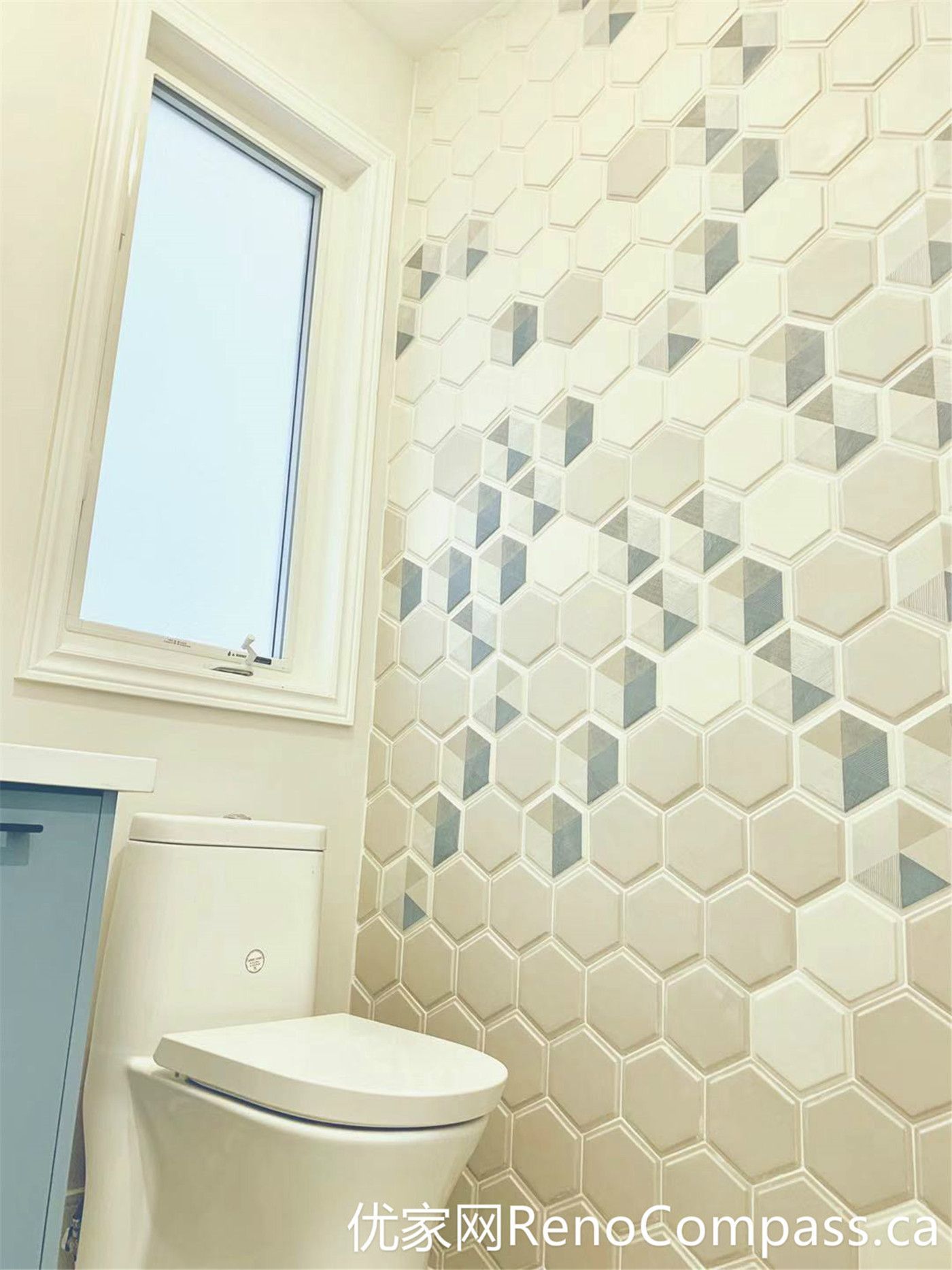 This Markham home renovation highlights a stylish bathroom featuring a soft blue vanity, round mirror, and a decorative hexagon tile accent wall. The combination of warm lighting, modern fixtures, and natural daylight from the window creates a cozy yet contemporary atmosphere with strong visual character.