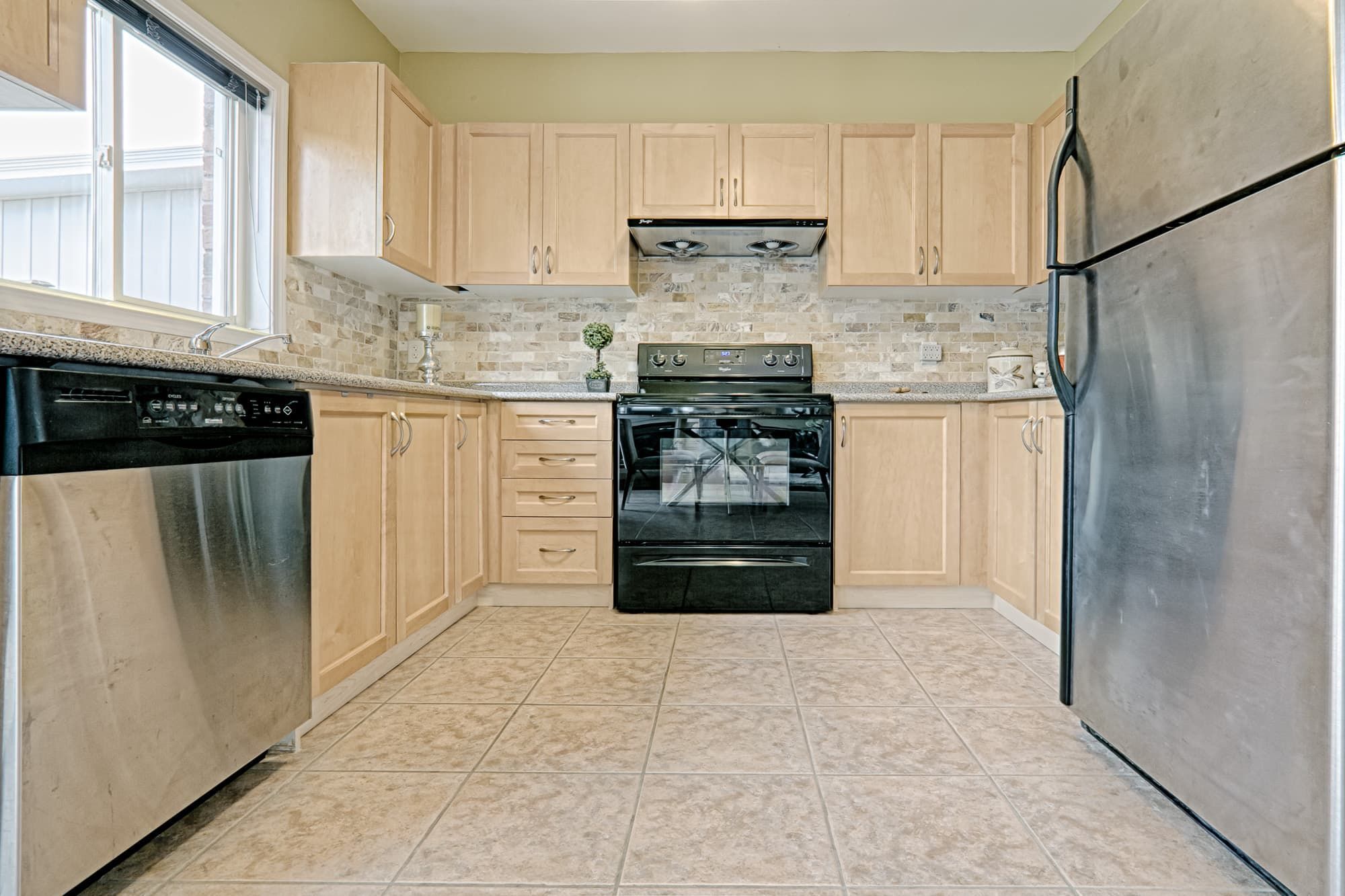 This bright kitchen features a simple and lovely interior design with light wood cabinetry, stainless steel appliances, and a neutral tile backsplash. The functional U-shaped layout maximizes storage and counter space, while large windows bring in natural light to enhance the overall brightness. This kitchen design focuses on practicality, clean lines, and everyday usability, making it ideal for family homes and rental properties.