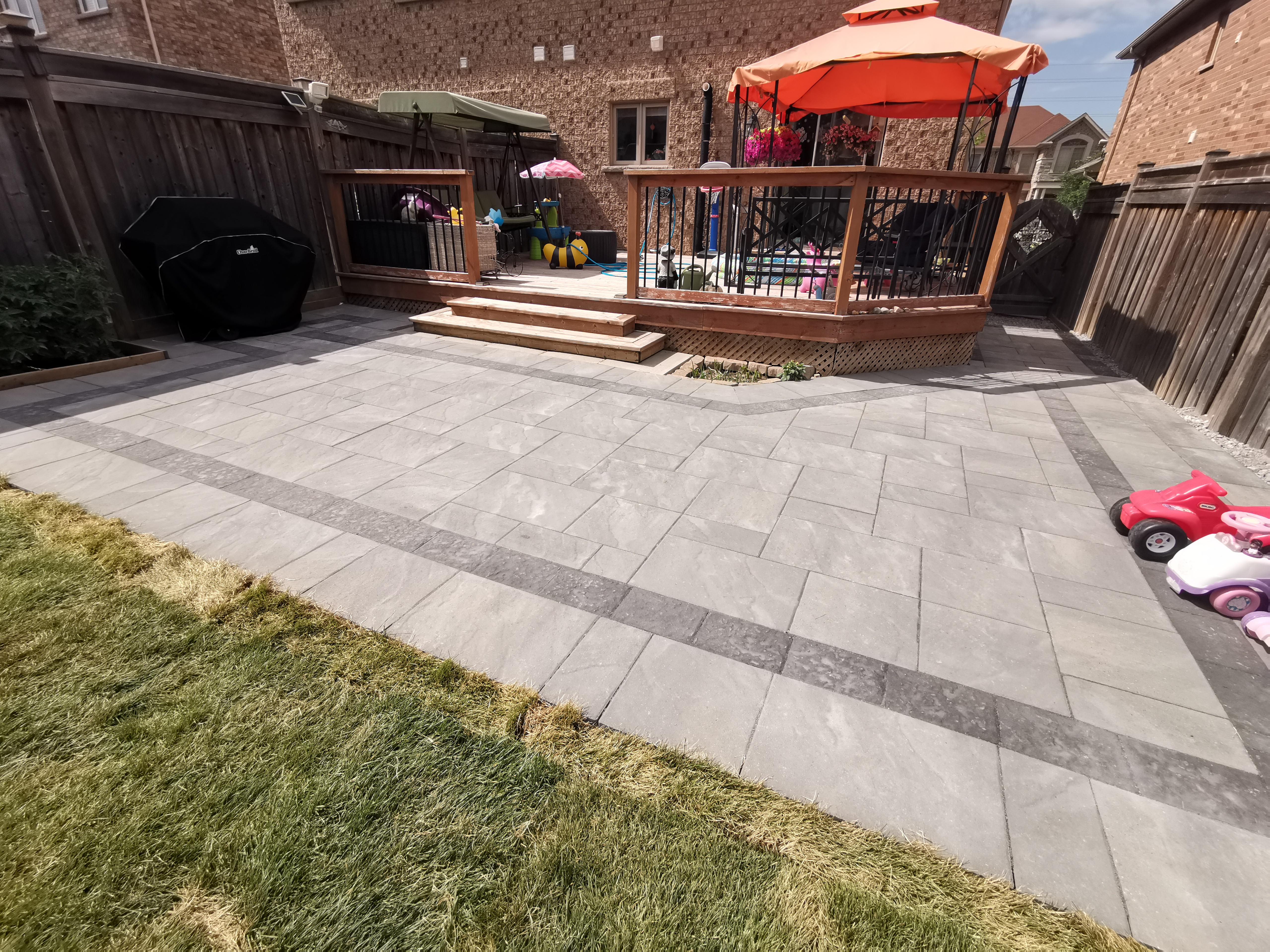 This Aurora backyard interlocking paver patio is designed to create a functional and inviting outdoor living space. Large-format concrete pavers are installed in a clean linear pattern with a contrasting border, enhancing visual definition and long-term durability. The patio seamlessly connects to a raised wooden deck with integrated steps, allowing smooth movement between indoor and outdoor areas. This professionally built interlock patio offers excellent drainage, stability, and low maintenance, making it ideal for family use and everyday enjoyment in residential backyards across Aurora.