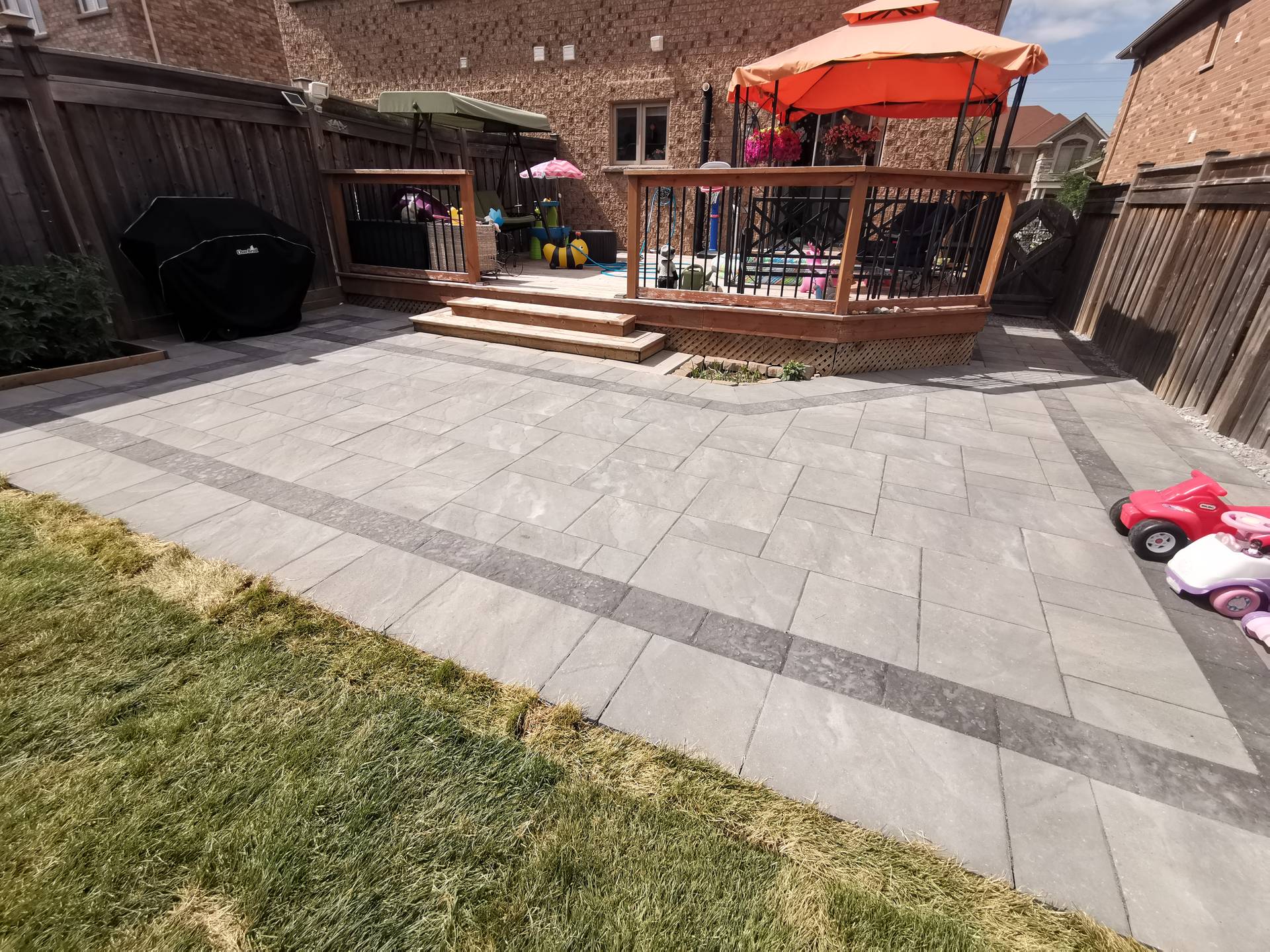 This Aurora backyard interlocking paver patio is designed to create a functional and inviting outdoor living space. Large-format concrete pavers are installed in a clean linear pattern with a contrasting border, enhancing visual definition and long-term durability. The patio seamlessly connects to a raised wooden deck with integrated steps, allowing smooth movement between indoor and outdoor areas. This professionally built interlock patio offers excellent drainage, stability, and low maintenance, making it ideal for family use and everyday enjoyment in residential backyards across Aurora.
