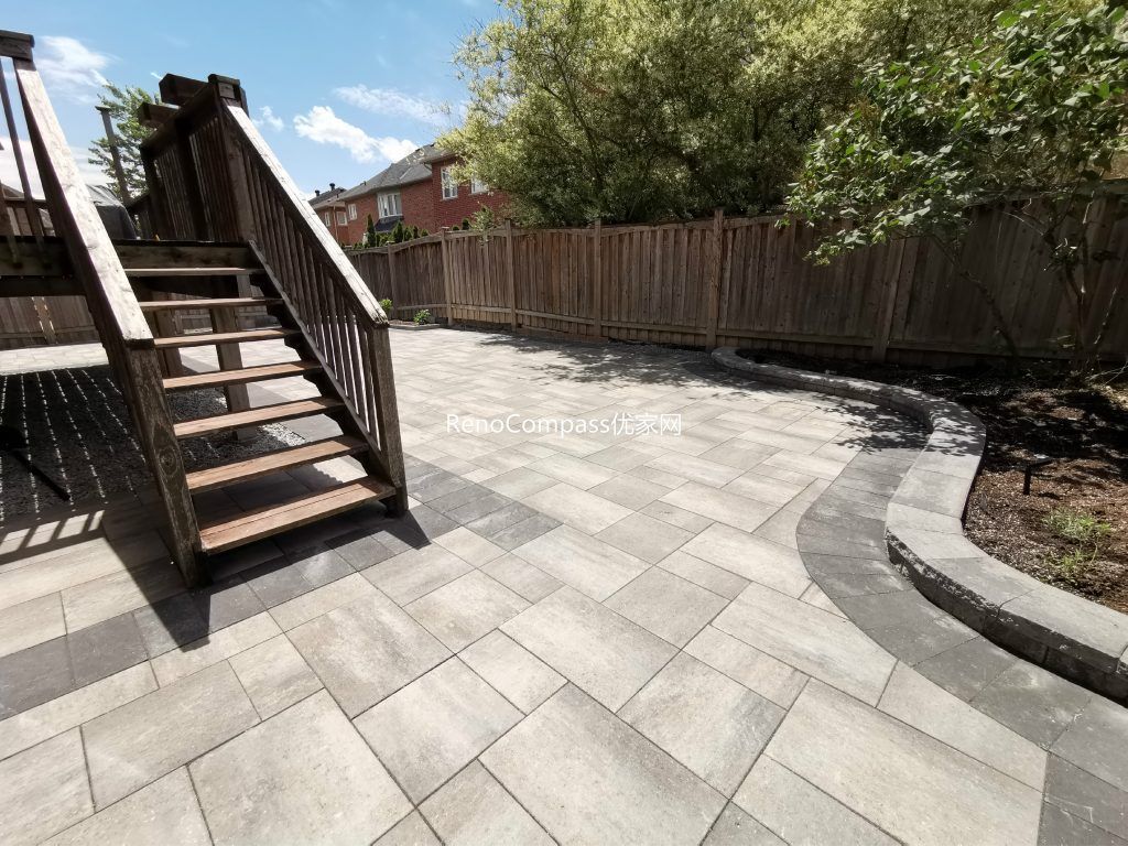 Modern backyard interlocking patio in Richmond Hill featuring elegant gray pavers and a curved stone border around the garden. The project seamlessly integrates with the wooden deck and stairs, creating a clean and functional outdoor space designed by RenoCompass for durability and style.