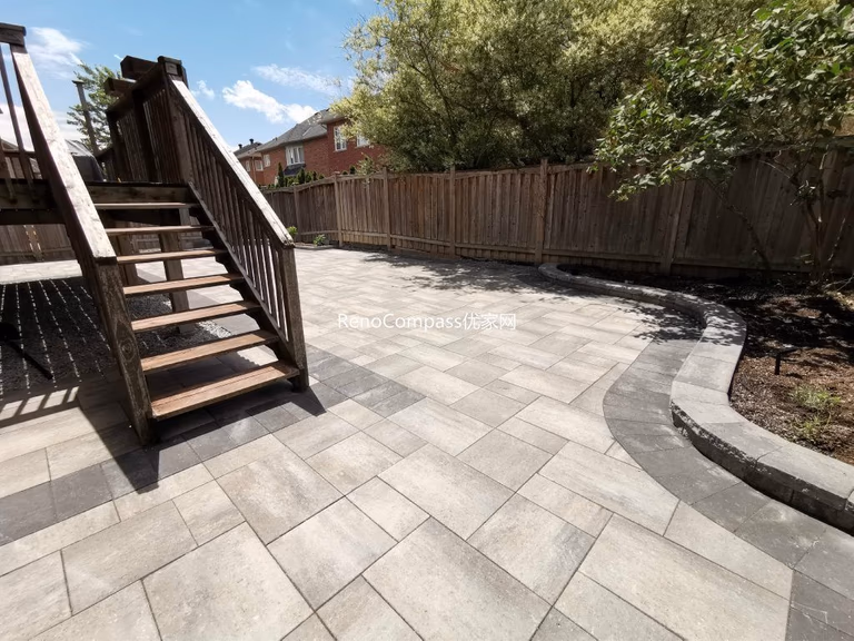 Modern backyard interlocking patio in Richmond Hill featuring elegant gray pavers and a curved stone border around the garden. The project seamlessly integrates with the wooden deck and stairs, creating a clean and functional outdoor space designed by RenoCompass for durability and style.