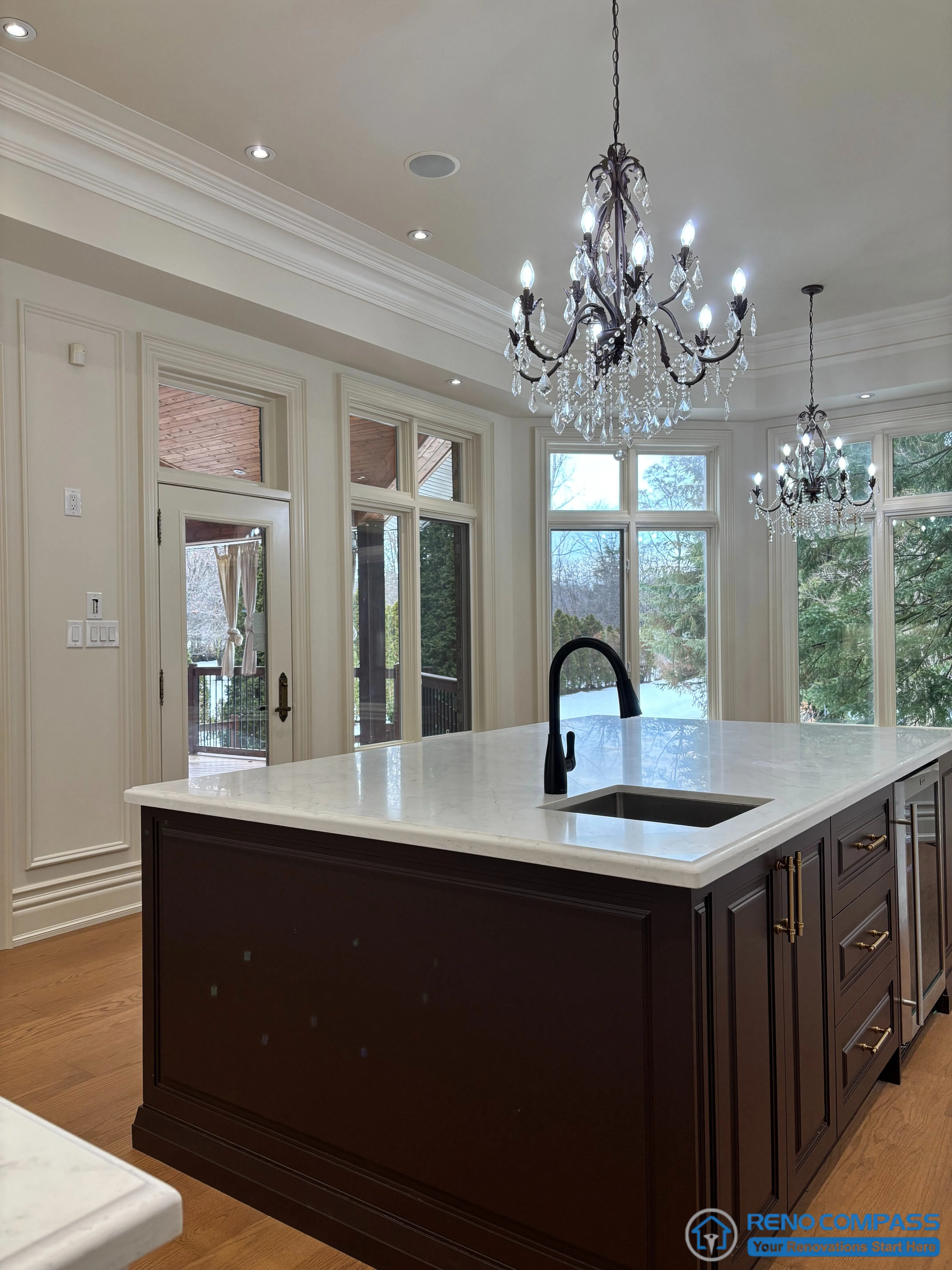 This European-style kitchen features a dark wood central island paired with a white stone countertop, creating a strong visual contrast rooted in classical proportion and symmetry. Detailed crown moulding, full-height windows, and a crystal chandelier reinforce the traditional European architectural language, while the island layout supports both daily cooking and family gathering. The design balances refined detailing with practical functionality, reflecting a timeless approach to high-end residential kitchen renovation in Richmond Hill.
