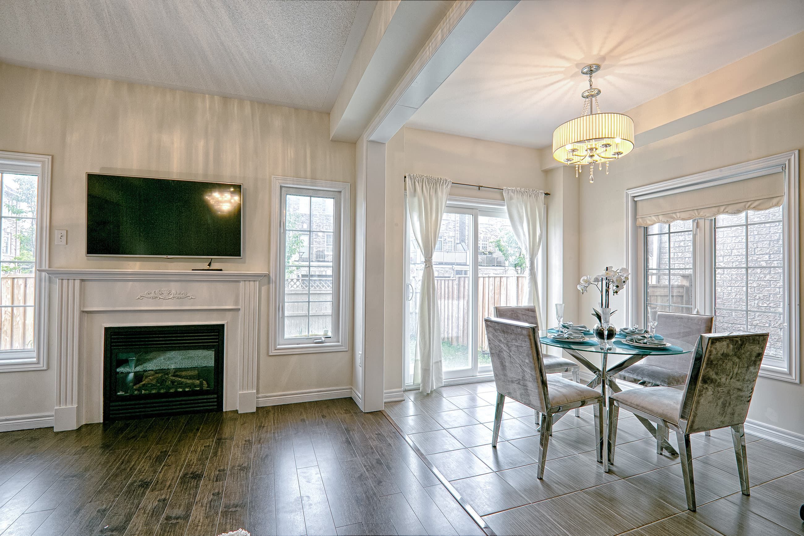 This open-concept dining and living space features a modern fireplace wall with mounted TV, elegant chandelier lighting, and large windows that bring in abundant natural light. The seamless transition between dark hardwood flooring and tiled dining space enhances visual flow and functionality. Designed with a balanced neutral palette and contemporary finishes, this interior reflects the craftsmanship of a professional Toronto renovation company specializing in stylish, functional, and modern home renovation projects.