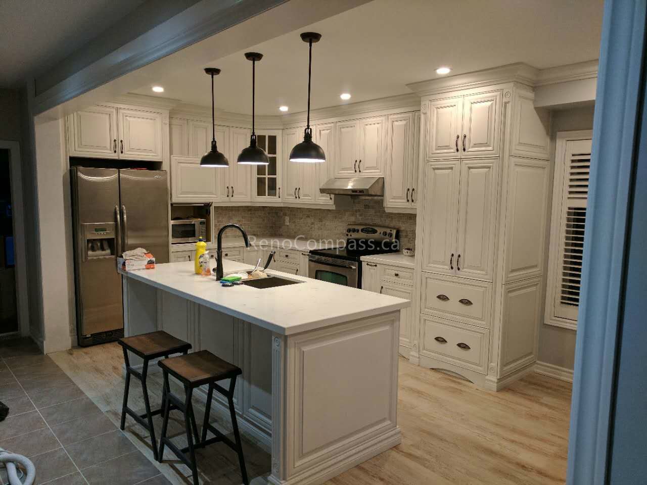 This completed Vaughan kitchen renovation by Reno Compass Inc. features custom white shaker cabinetry, a spacious quartz waterfall island with seating, and stylish black pendant lighting. The redesigned layout improves storage and functionality while stainless steel appliances and warm wood flooring create a bright, modern, and inviting kitchen space perfect for everyday family living and entertaining.