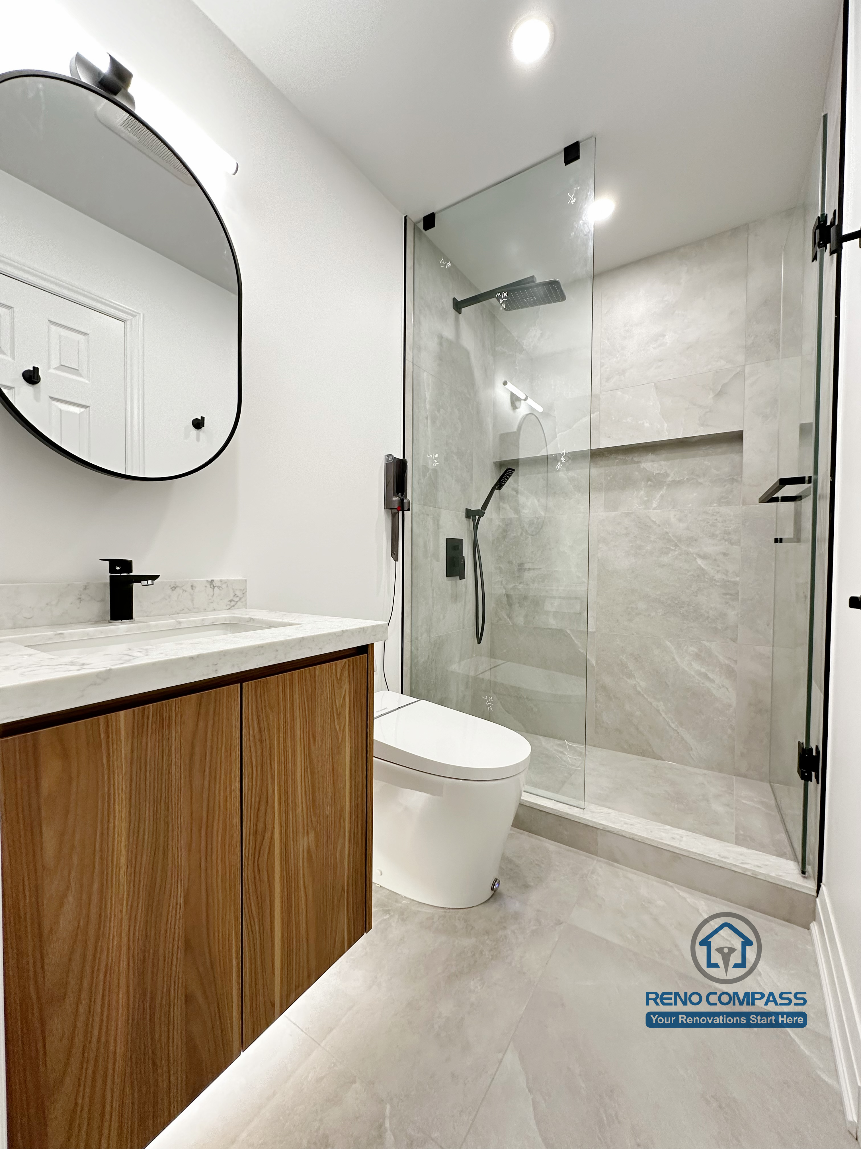 Bathroom renovation in Markham featuring a custom wood vanity with quartz countertop, round wall-mounted mirror, and a frameless glass shower with built-in niche. Clean lines, neutral finishes, and modern fixtures create a bright and functional bathroom layout.