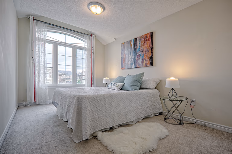 This modern master bedroom renovation showcases a calm and elegant design with soft neutral tones, plush carpeting, and large arched windows that bring in abundant natural light. The clean layout, cozy bedding, and contemporary bedside lighting create a relaxing and comfortable atmosphere ideal for everyday living. Completed by a professional Toronto renovation company, this bedroom upgrade highlights thoughtful space planning and refined interior finishes tailored for modern residential homes.