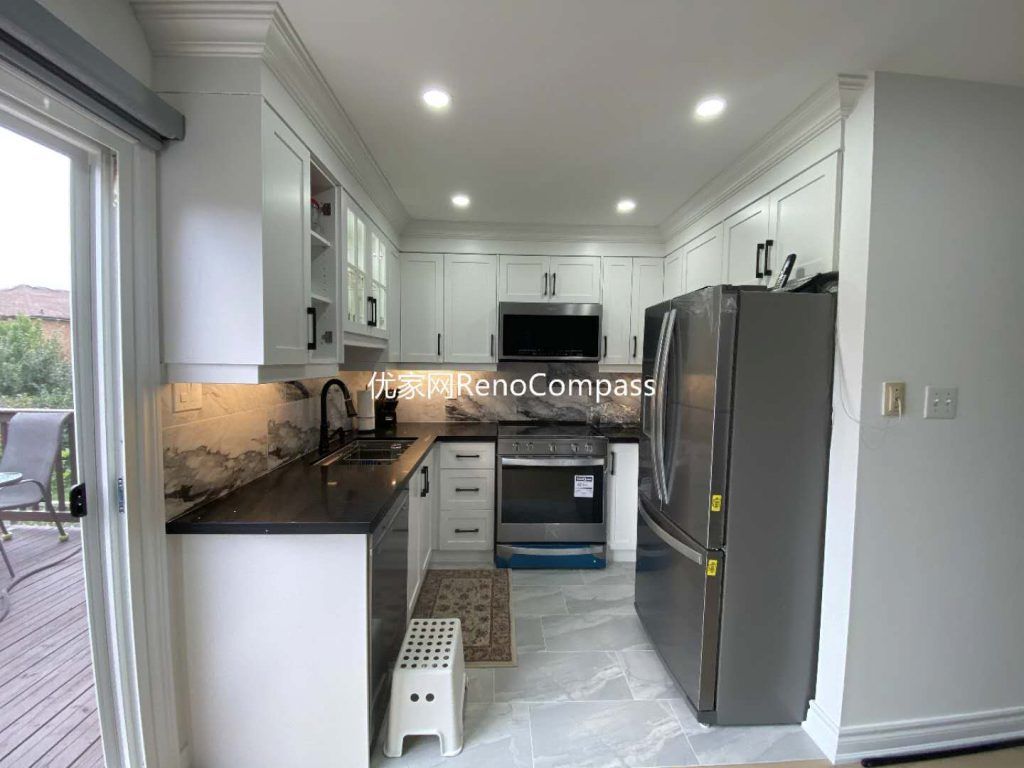 This image presents an additional view of a renovated kitchen in Mississauga, highlighting the efficient layout, custom white cabinetry, and improved natural light connection. Upper glass-front cabinets add visual openness and practical display storage, while under-cabinet lighting enhances task visibility along the stone-pattern backsplash. The combination of quartz countertops, stainless steel appliances, and durable tile flooring creates a balanced kitchen designed for daily use. Large sliding doors provide natural light and outdoor access, reinforcing the functional and comfortable design approach delivered by a professional Toronto renovation company.