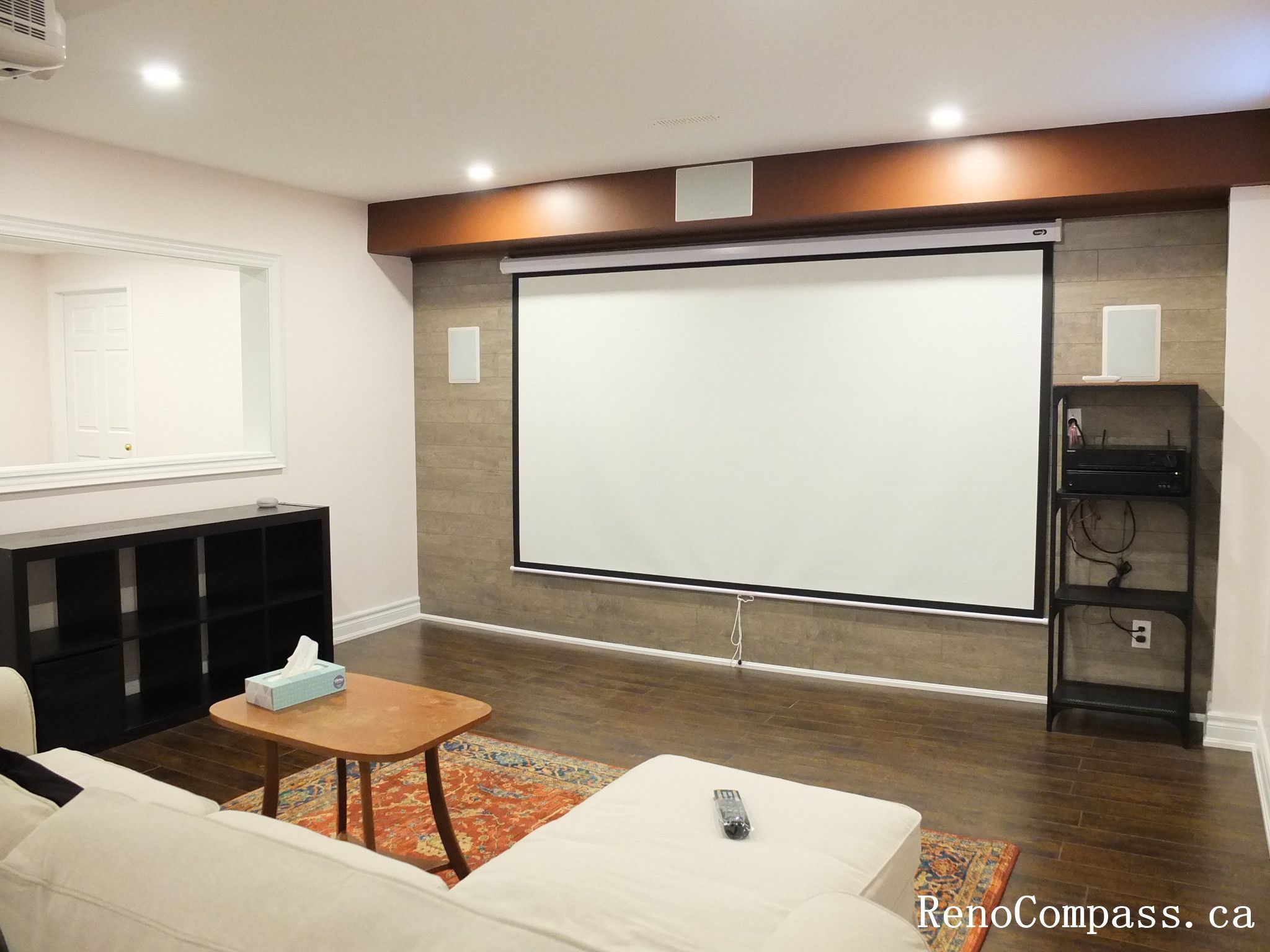 This Newmarket basement renovation features a dedicated home theater area designed for comfortable movie nights and entertainment. The space includes a large projection screen, recessed pot lighting, dark hardwood flooring, and a feature wall with integrated audio speakers for an immersive viewing experience. A cozy seating area with modern furniture and a warm accent beam creates a welcoming atmosphere while maintaining a clean and contemporary design. Completed by Reno Compass Inc., this project showcases a stylish and functional basement entertainment space tailored for modern family living.