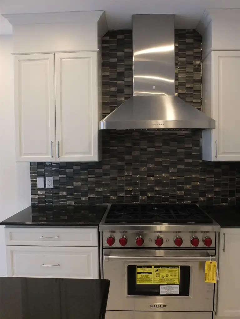 This Kleinburg interior renovation showcases a modern kitchen featuring custom white cabinetry, a spacious island with integrated sink, and sleek black countertops. The kitchen is highlighted by a professional stainless steel gas range and hood, complemented by a dark mosaic tile backsplash that adds depth and contrast. Elegant pendant lighting and large windows enhance the bright, open atmosphere, creating a refined and functional cooking space ideal for modern living.