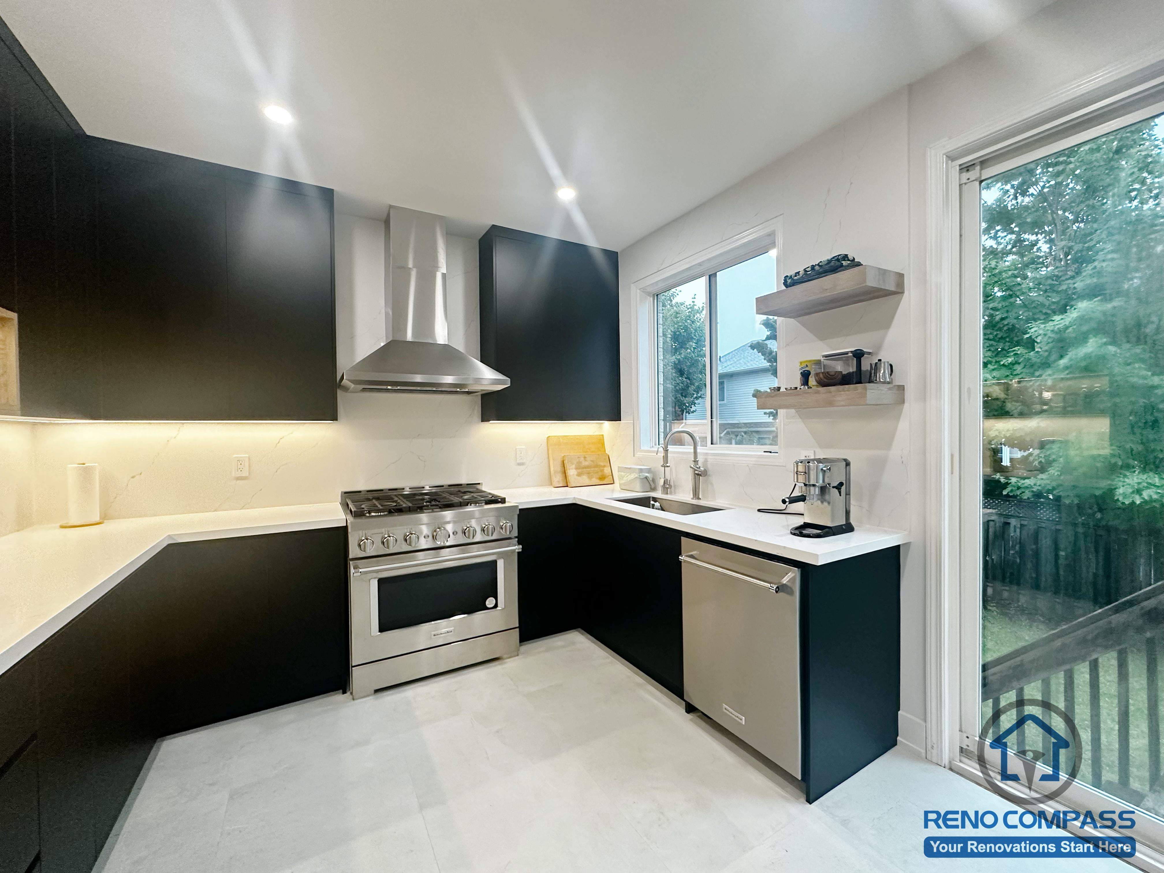 This contemporary kitchen renovation in Oakville features black cabinetry paired with white countertops and a stainless steel gas range as the focal point. Under-cabinet lighting enhances task areas, while open shelving and a large window bring in natural light and everyday functionality. The U-shaped layout supports efficient movement and a clean, practical cooking environment designed for daily use.