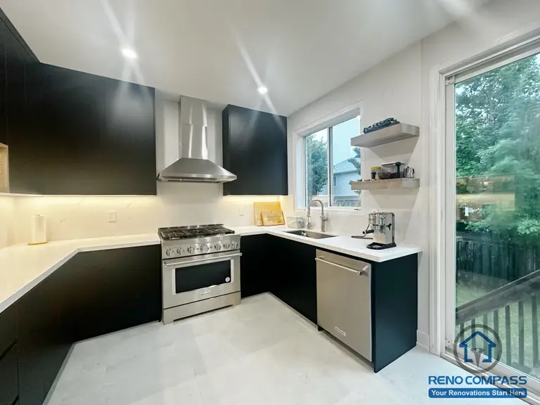 This contemporary kitchen renovation in Oakville features black cabinetry paired with white countertops and a stainless steel gas range as the focal point. Under-cabinet lighting enhances task areas, while open shelving and a large window bring in natural light and everyday functionality. The U-shaped layout supports efficient movement and a clean, practical cooking environment designed for daily use.