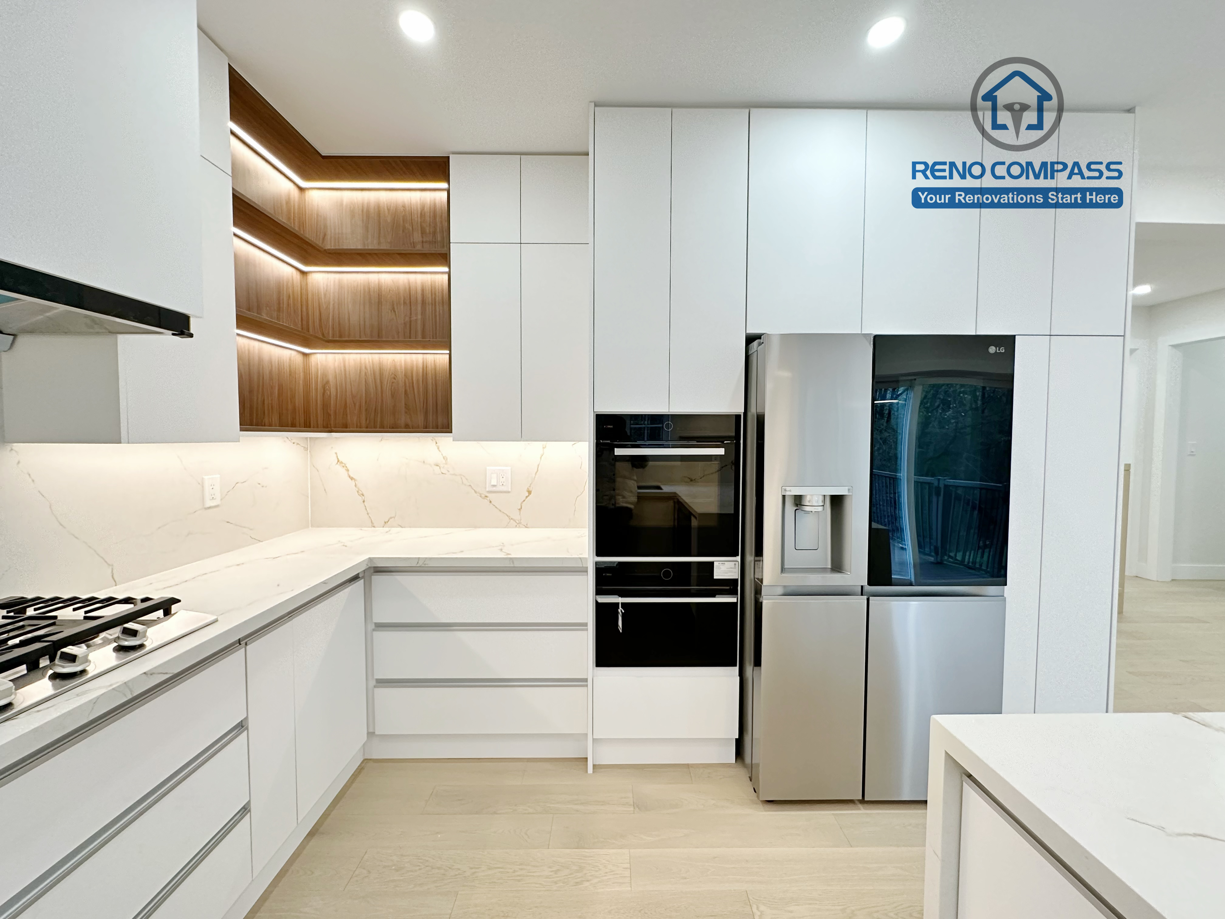 This image highlights a modern kitchen renovation in Richmond Hill featuring full-height custom white cabinetry with integrated built-in appliances, including a stainless steel refrigerator and wall oven. The design incorporates seamless storage solutions, quartz countertops, and warm wood accent shelving with integrated LED lighting for added depth and visual interest. Recessed ceiling lighting and light hardwood flooring complete the clean, contemporary aesthetic. Completed by a professional Toronto Renovation Company, this kitchen renovation demonstrates efficient space planning, premium materials, and refined craftsmanship.