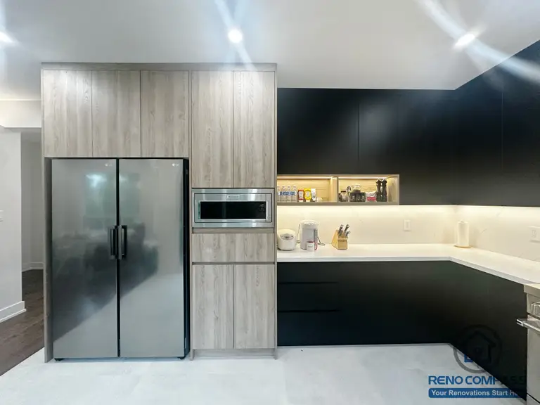 This Oakville interior renovation presents a modern kitchen designed with integrated wood-grain cabinetry and matte black lower storage for a clean, structured appearance. Built-in appliances are seamlessly incorporated into the cabinet wall, maximizing storage efficiency while maintaining a streamlined look. Under-cabinet lighting enhances both functionality and depth along the countertop workspace. The combination of contrasting finishes and minimal detailing emphasizes clarity, balance, and long-term usability, making this kitchen well suited for everyday cooking and modern family living.