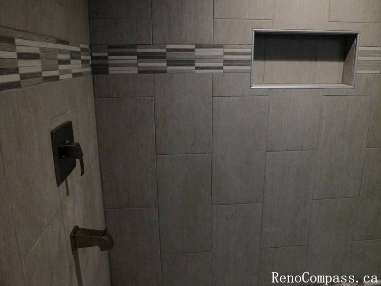 Close‑up view of a modern tiled shower wall featuring a horizontal accent strip, built‑in storage niche, and matte black fixtures. A stylish upgrade in this Waterloo basement bathroom renovation