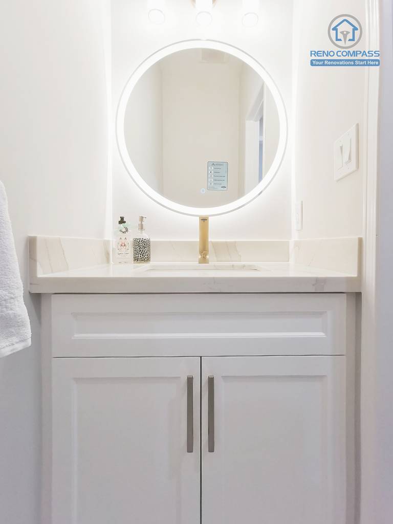 This Markham interior renovation features a refined bathroom vanity designed with a clean, understated aesthetic and practical daily use in mind. The custom white shaker vanity provides concealed storage while maintaining a balanced, symmetrical appearance. A quartz countertop offers durability and easy maintenance, paired with a warm-toned faucet that adds subtle contrast. The circular LED mirror introduces soft, even illumination, enhancing visibility without harsh glare and contributing to a calm, spa-like atmosphere. Completed by Reno Compass, this bathroom renovation reflects careful material coordination, proportion control, and a timeless design approach tailored for modern Markham homes.