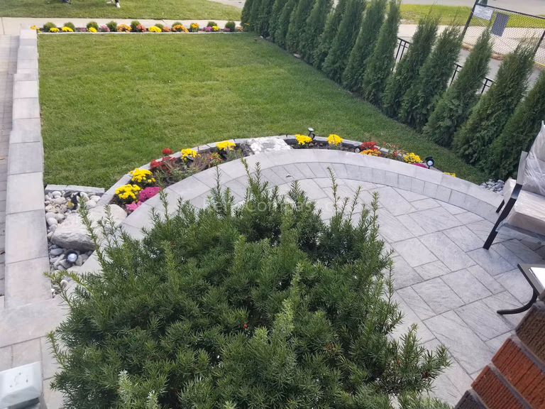 This Toronto landscaping renovation showcases a beautifully crafted curved stone patio with integrated garden beds and a spacious green lawn. The interlock patio design creates a functional outdoor seating area, while raised flower beds and privacy cedar hedges enhance structure and visual appeal. Natural stone details and soft landscape lighting complete the space, delivering a modern, low-maintenance backyard landscaping upgrade by a professional Toronto landscaping renovation team.