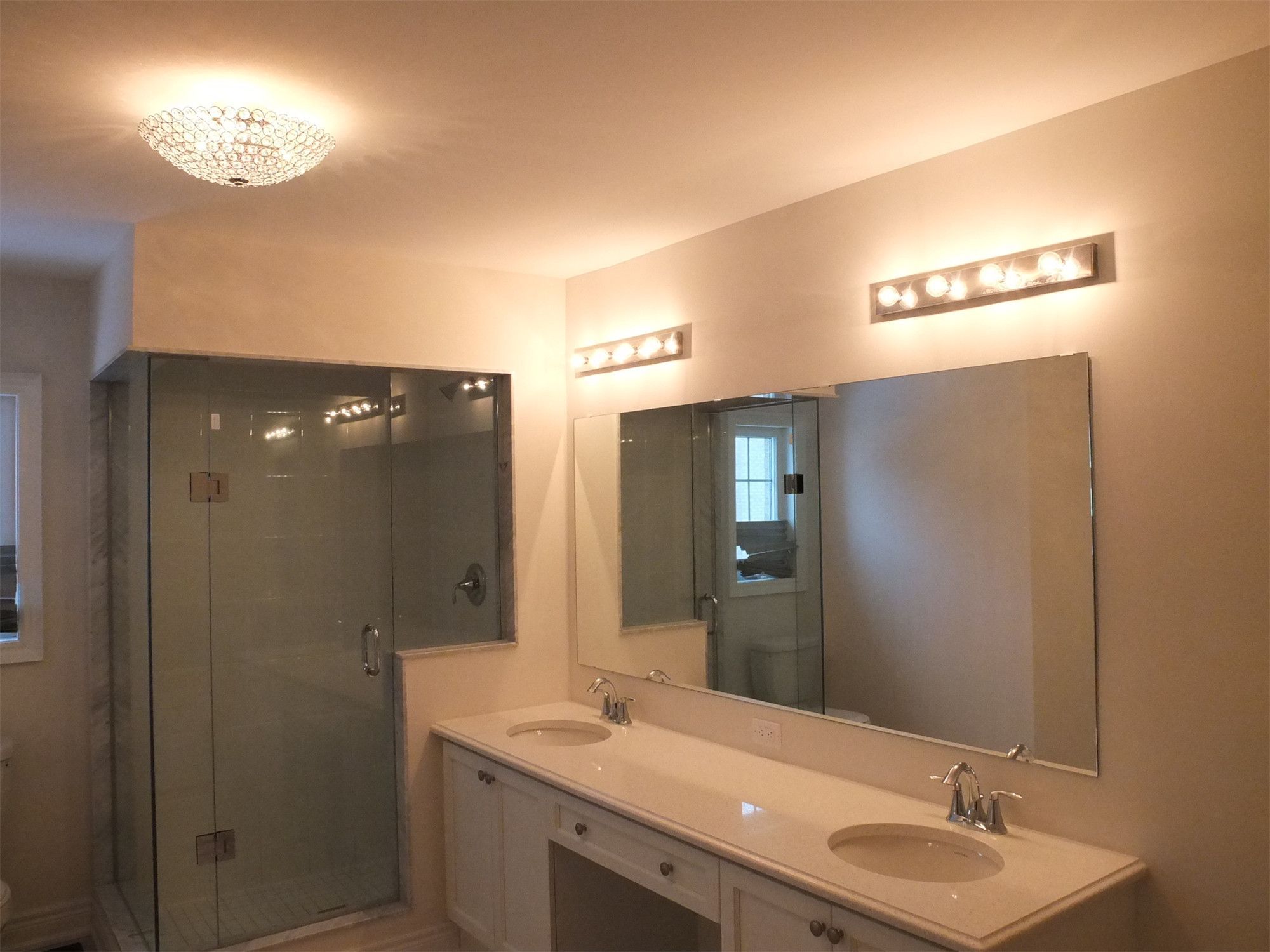 This Kleinburg interior renovation features a bright and modern bathroom with a spacious double vanity, large wall mirror, and elegant lighting fixtures. The design includes a frameless glass shower enclosure, contemporary faucets, and clean white cabinetry that enhances the sense of space and brightness. Soft ceiling lighting and vanity lights provide balanced illumination, creating a comfortable and refined bathroom environment.