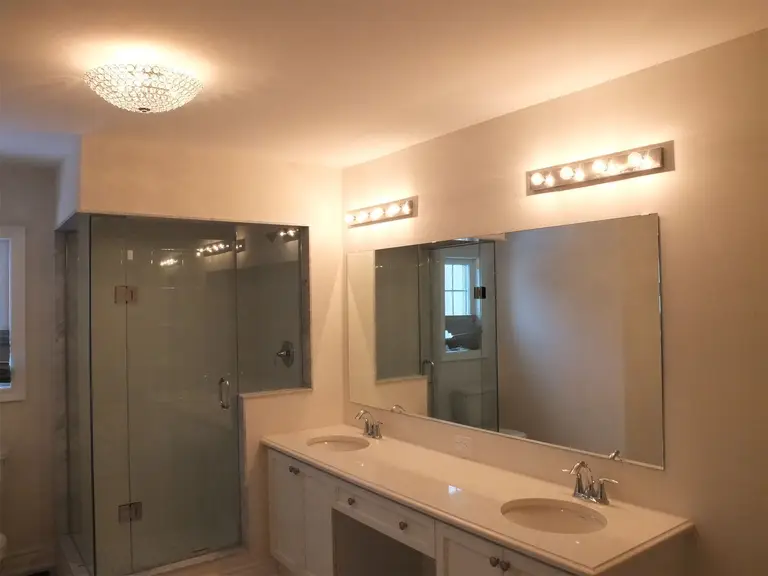 This Kleinburg interior renovation features a bright and modern bathroom with a spacious double vanity, large wall mirror, and elegant lighting fixtures. The design includes a frameless glass shower enclosure, contemporary faucets, and clean white cabinetry that enhances the sense of space and brightness. Soft ceiling lighting and vanity lights provide balanced illumination, creating a comfortable and refined bathroom environment.