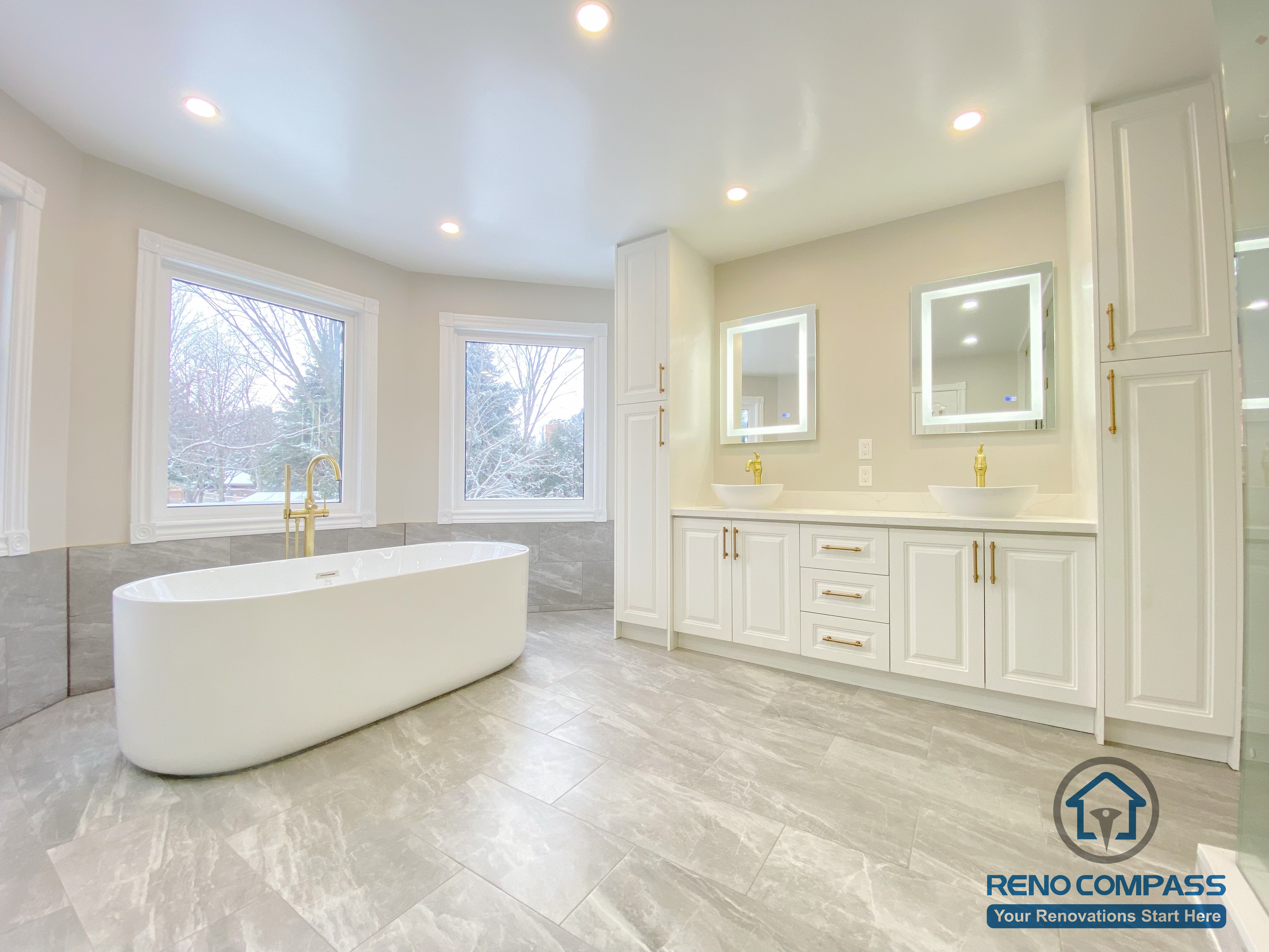 This Richmond Hill bathroom renovation highlights a modern freestanding bathtub positioned in front of large bay windows, creating a bright and relaxing spa-like atmosphere. The space features large-format grey floor tiles, marble-style wall finishes, recessed ceiling lighting, and gold-finish tub fixtures. The clean layout and natural light enhance comfort, elegance, and functionality, showcasing a thoughtfully designed luxury bathroom renovation in Richmond Hill.