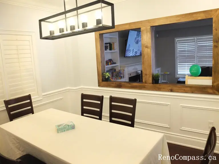 This Newmarket interior renovation highlights a bright and elegant dining room designed for both everyday meals and family gatherings. The space features classic wall wainscoting, dark hardwood flooring, and a modern rectangular pendant light fixture that adds a contemporary touch above the dining table. A large interior window opening with a warm wood frame connects the dining area to the adjacent living space, creating a more open and interactive layout. Completed by Reno Compass Inc., this project demonstrates thoughtful interior design and quality craftsmanship for modern family homes.