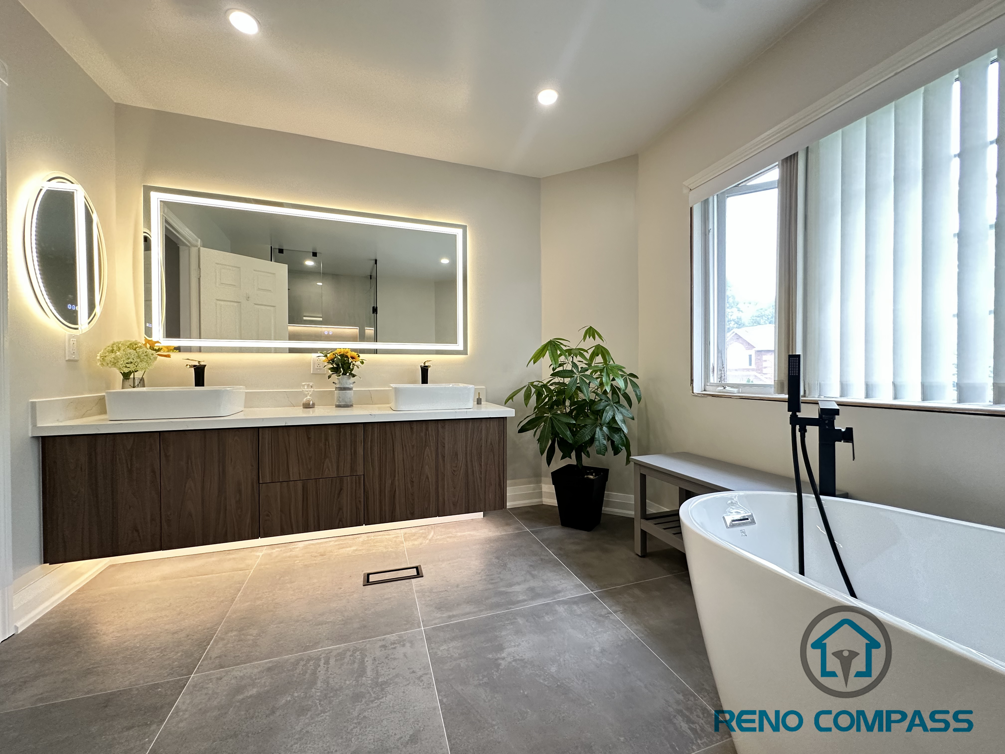 A spacious Oakville bathroom renovation featuring a floating double vanity topped with quartz, a stylish round LED mirror, large-format floor tiles, and a freestanding bathtub. This design combines modern elegance with functional luxury, creating a serene and cohesive spa-like retreat.