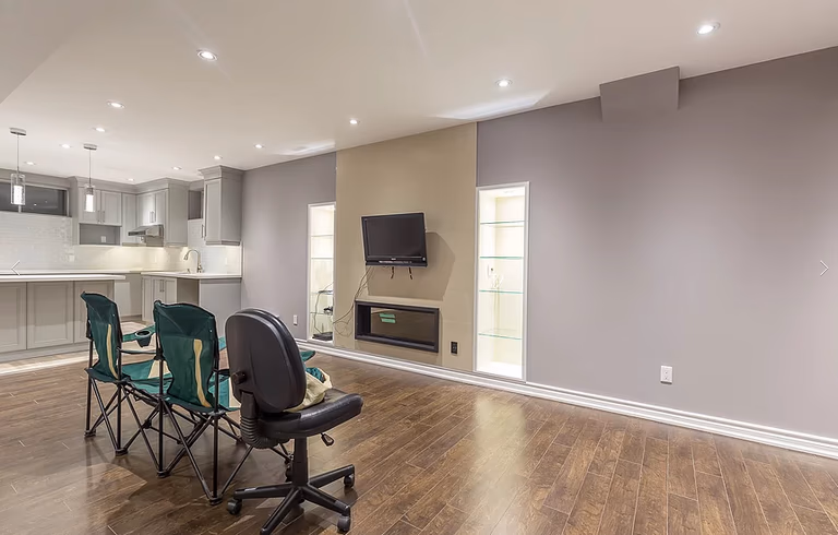 This Toronto basement renovation features a contemporary open-concept layout connecting the basement kitchen and living area. The space includes engineered hardwood flooring, recessed LED pot lights, a wall-mounted electric fireplace with custom niche lighting, and a modern grey and beige accent wall design. The seamless integration of kitchen and entertainment space enhances comfort and functionality, professionally completed by Reno Compass Inc., a leading Toronto renovation company.