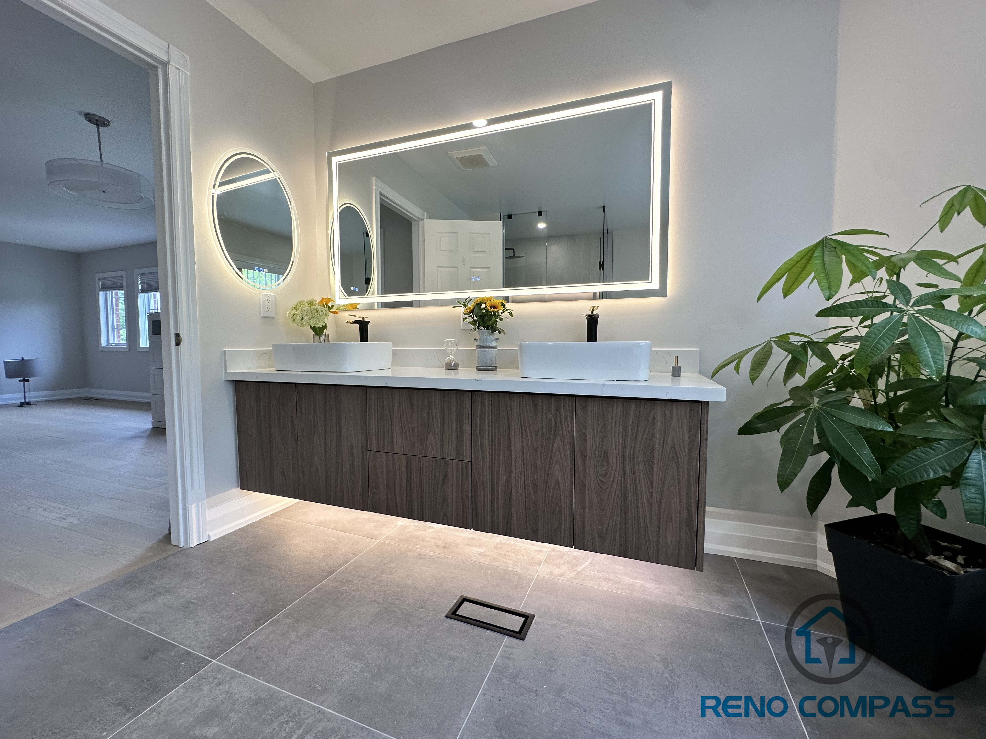 spacious Oakville bathroom renovation featuring a floating double vanity topped with quartz, a stylish round LED mirror, large-format floor tiles, and a freestanding bathtub. This design combines modern elegance with functional luxury, creating a serene and cohesive spa-like retreat.