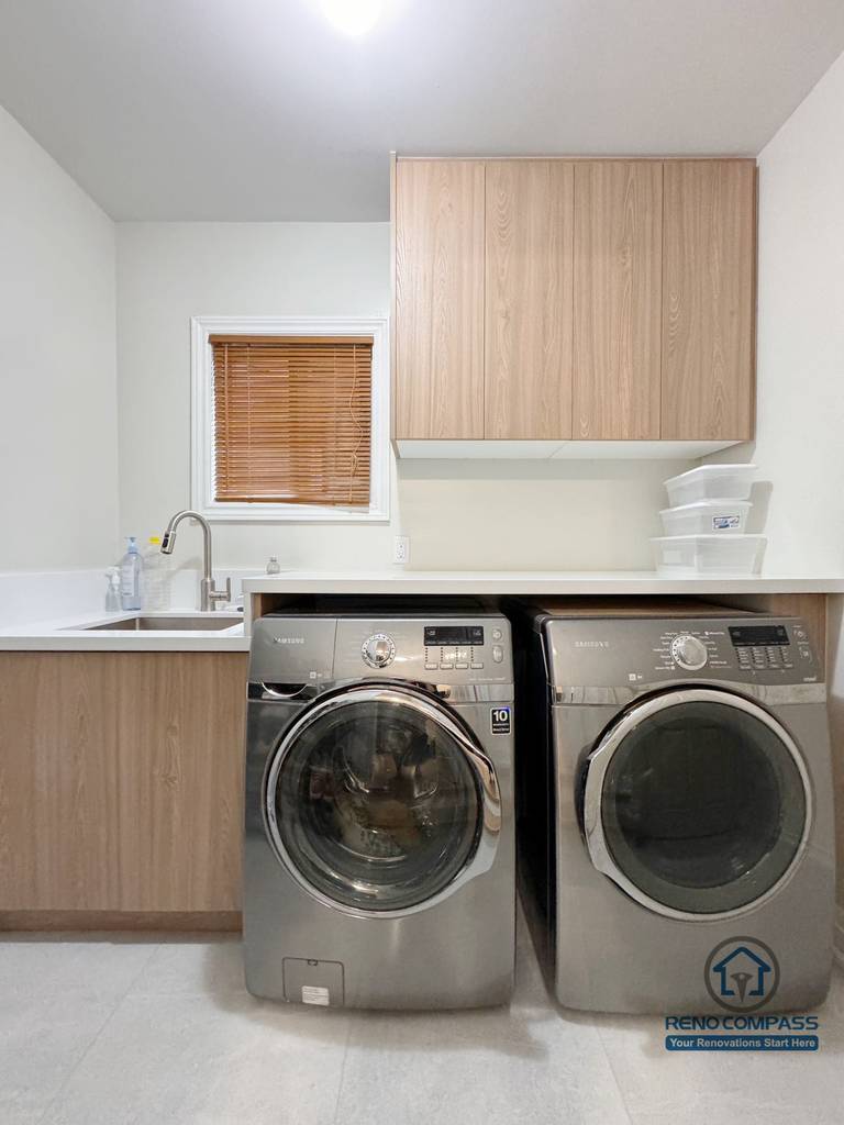 This Mississauga interior renovation showcases a modern laundry room designed for both efficiency and clean aesthetics. Custom wood cabinetry provides ample storage, while the integrated countertop and sink create a practical workspace for daily use. The side-by-side washer and dryer layout maximizes functionality, resulting in a bright, organized, and contemporary laundry area.