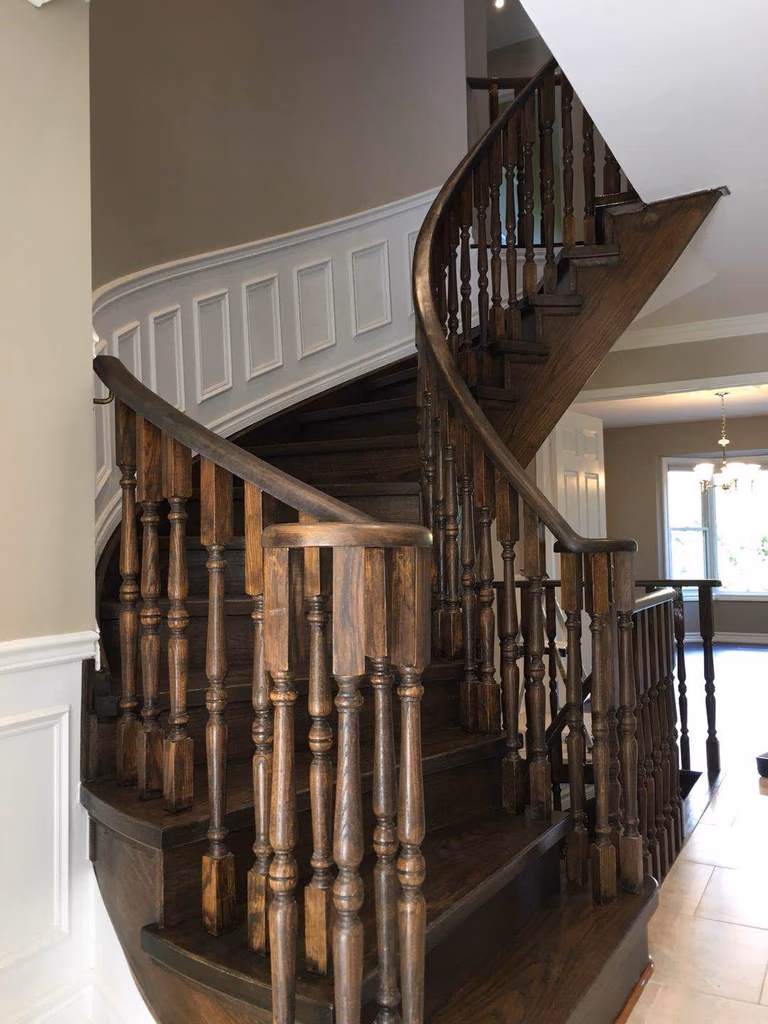 This Toronto interior renovation showcases an elegant curved hardwood staircase featuring rich dark wood treads, detailed wooden balusters, and a smooth custom handrail. The staircase upgrade enhances both architectural character and structural durability while complementing the home’s interior design. Decorative wall paneling and neutral wall tones further highlight the craftsmanship, creating a refined focal point commonly seen in high-end Toronto residential interior renovations.