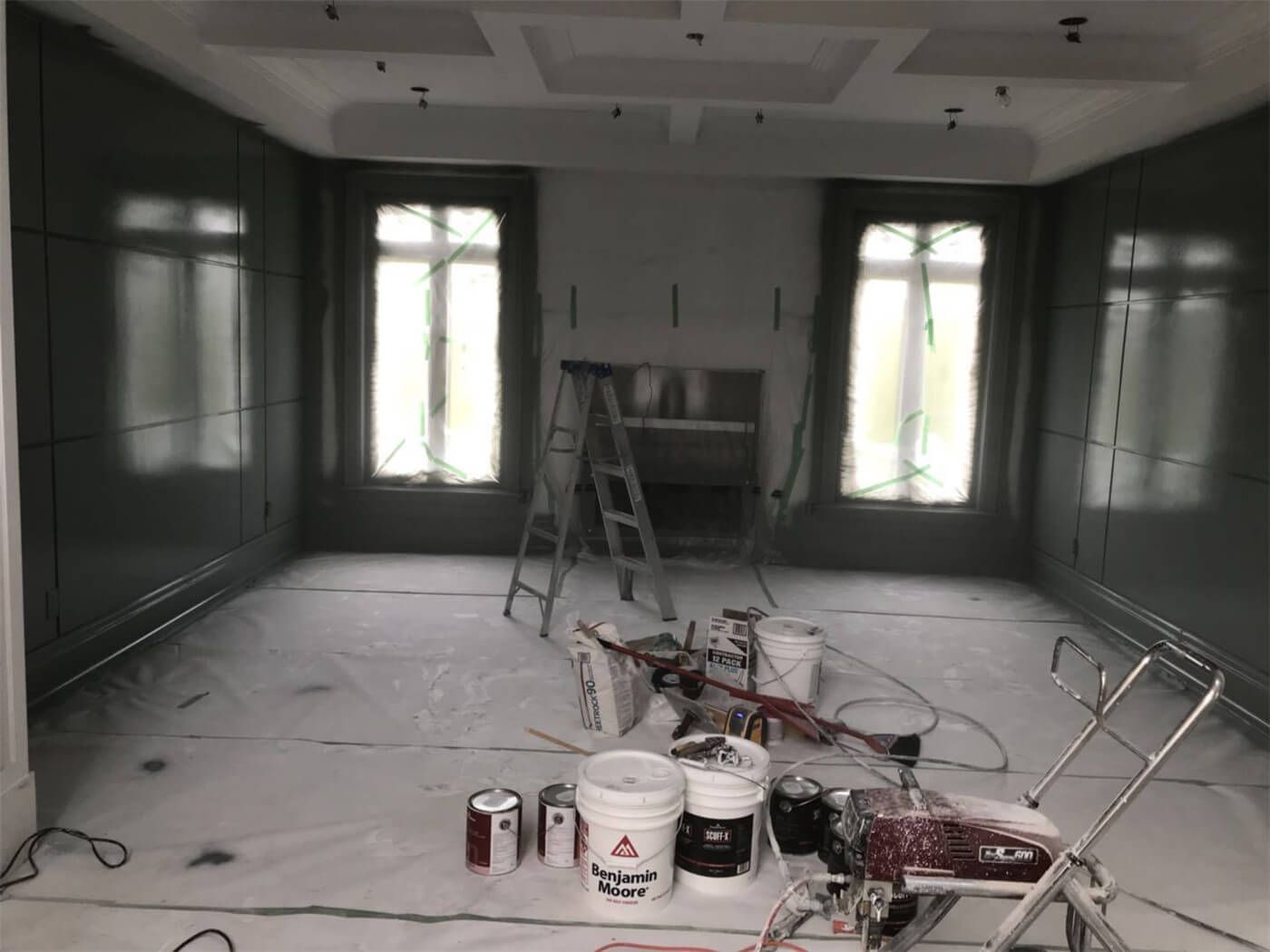 This image highlights a luxury interior wall painting project in Toronto, featuring custom wall paneling and a detailed coffered ceiling design. The space is carefully prepared with full floor protection, professional spray equipment, and premium paint materials to achieve a rich, smooth finish. Large windows and a central fireplace create a balanced focal point, while deep-toned walls add elegance and depth to the room. Our Toronto professional painting team delivers meticulous craftsmanship, precise edge work, and high-end interior finishes tailored to upscale residential homes.