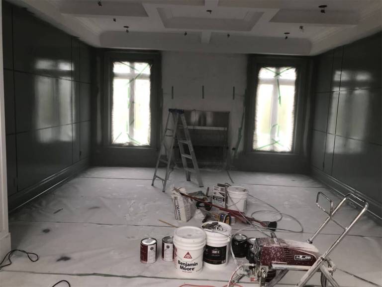 This image highlights a luxury interior wall painting project in Toronto, featuring custom wall paneling and a detailed coffered ceiling design. The space is carefully prepared with full floor protection, professional spray equipment, and premium paint materials to achieve a rich, smooth finish. Large windows and a central fireplace create a balanced focal point, while deep-toned walls add elegance and depth to the room. Our Toronto professional painting team delivers meticulous craftsmanship, precise edge work, and high-end interior finishes tailored to upscale residential homes.