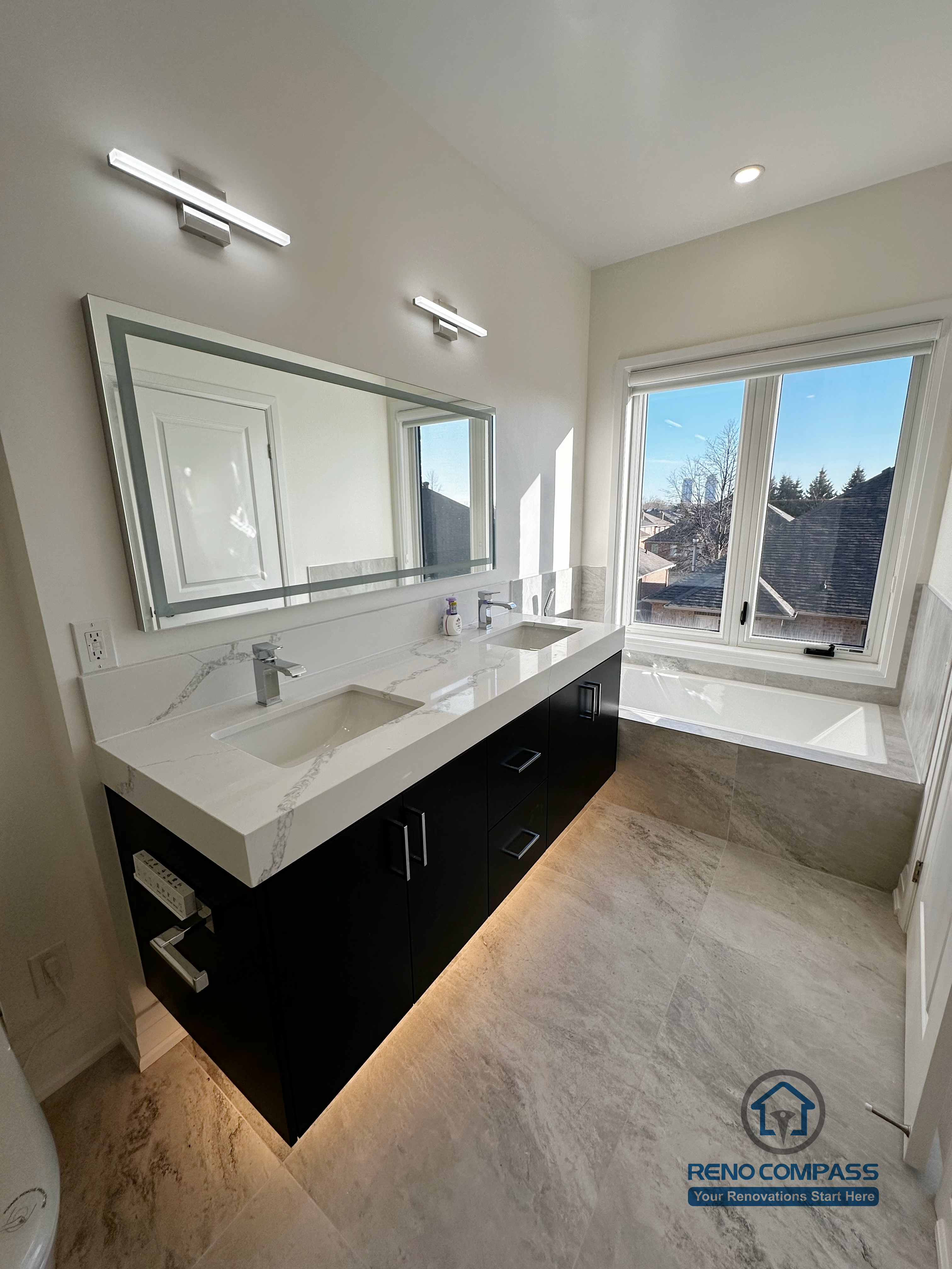 This image showcases a modern bathroom vanity completed as part of a Richmond Hill interior renovation. The design features a sleek quartz countertop with an integrated sink, custom dark-tone cabinetry with under-cabinet lighting, and a wide framed mirror paired with wall-mounted lighting. Natural light from the window enhances the clean layout and creates a bright, comfortable daily-use space.