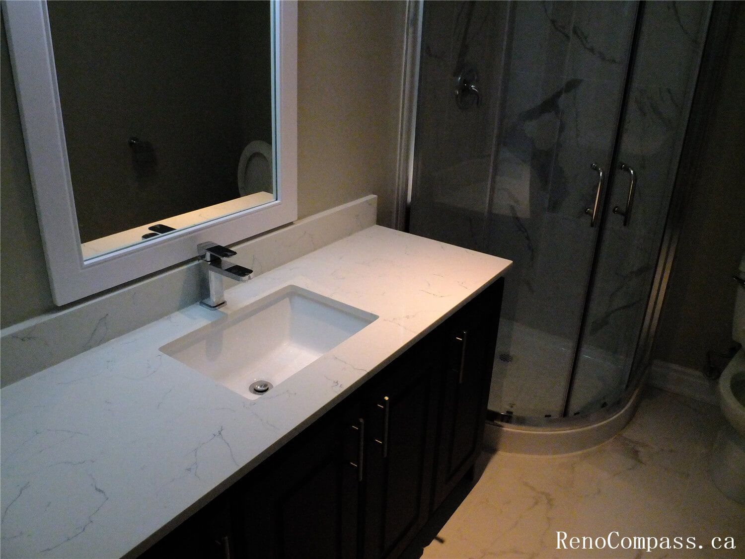 Close-up view of the bathroom vanity installed in this Ajax basement renovation, featuring a sleek quartz countertop with an integrated sink, modern black faucet, and dark storage cabinetry. The large mirror enhances brightness while the curved glass shower enclosure complements the contemporary bathroom design. Completed by a professional Toronto Renovation Company, this basement bathroom upgrade combines elegant finishes with practical functionality.