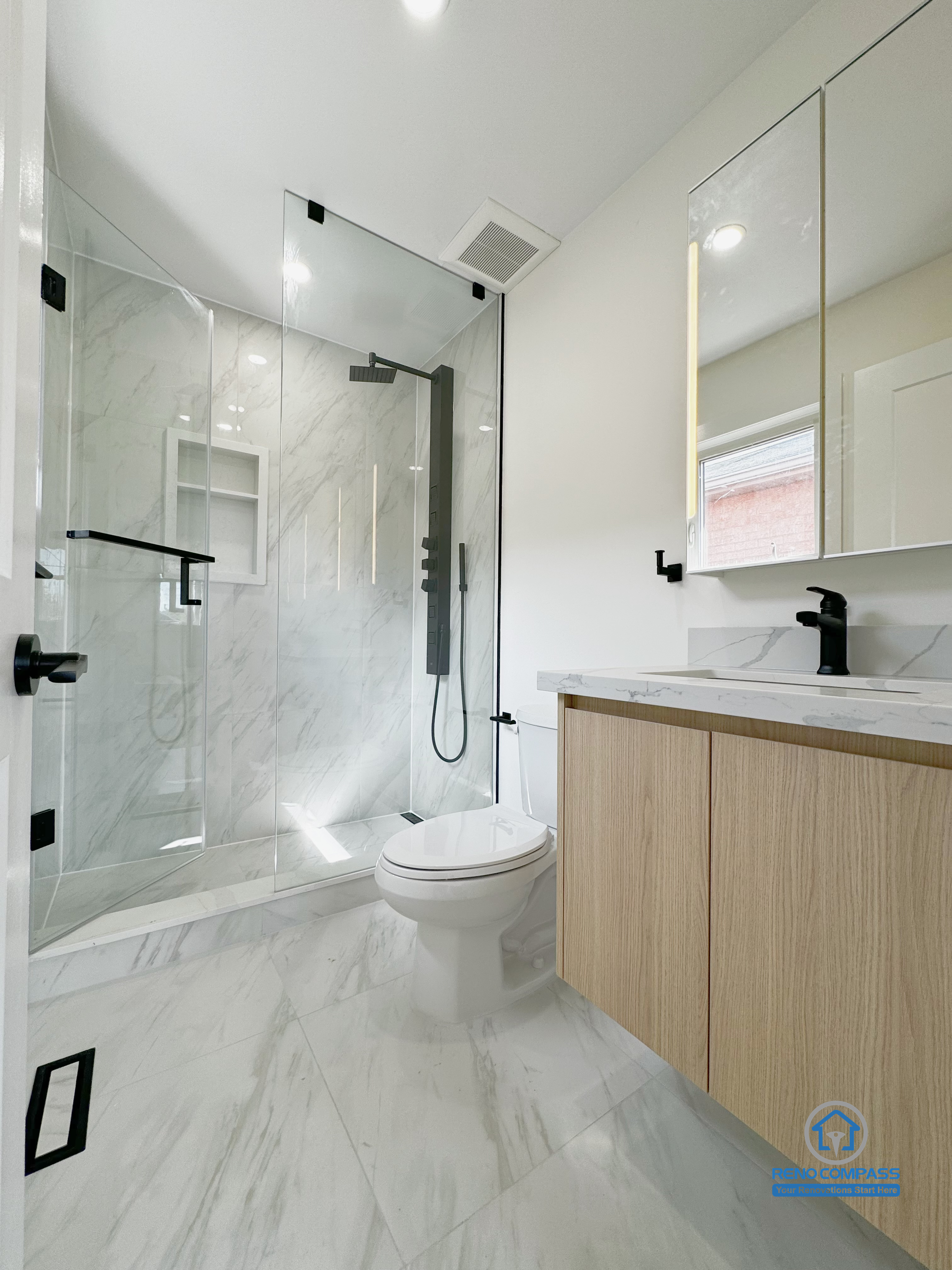 This image features a completed modern bathroom renovation in Richmond Hill, designed with a clean and functional layout. The space includes a full glass walk-in shower with marble-style wall tiles, matte black fixtures, and a streamlined drainage detail. A wood vanity adds warmth and contrast to the bright finishes, while integrated lighting and minimal hardware maintain a balanced, contemporary look. The design focuses on durability, daily comfort, and long-term residential use, making the bathroom both practical and visually cohesive.