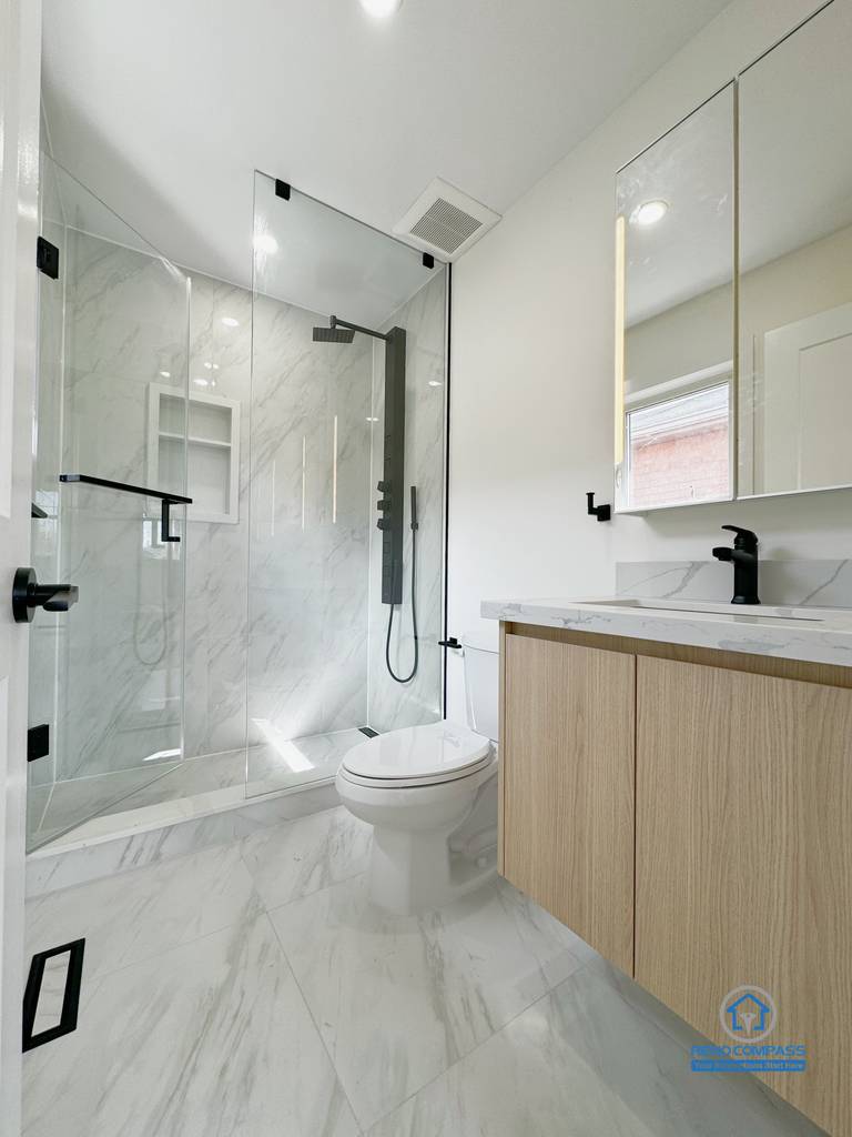 This image features a completed modern bathroom renovation in Richmond Hill, designed with a clean and functional layout. The space includes a full glass walk-in shower with marble-style wall tiles, matte black fixtures, and a streamlined drainage detail. A wood vanity adds warmth and contrast to the bright finishes, while integrated lighting and minimal hardware maintain a balanced, contemporary look. The design focuses on durability, daily comfort, and long-term residential use, making the bathroom both practical and visually cohesive.