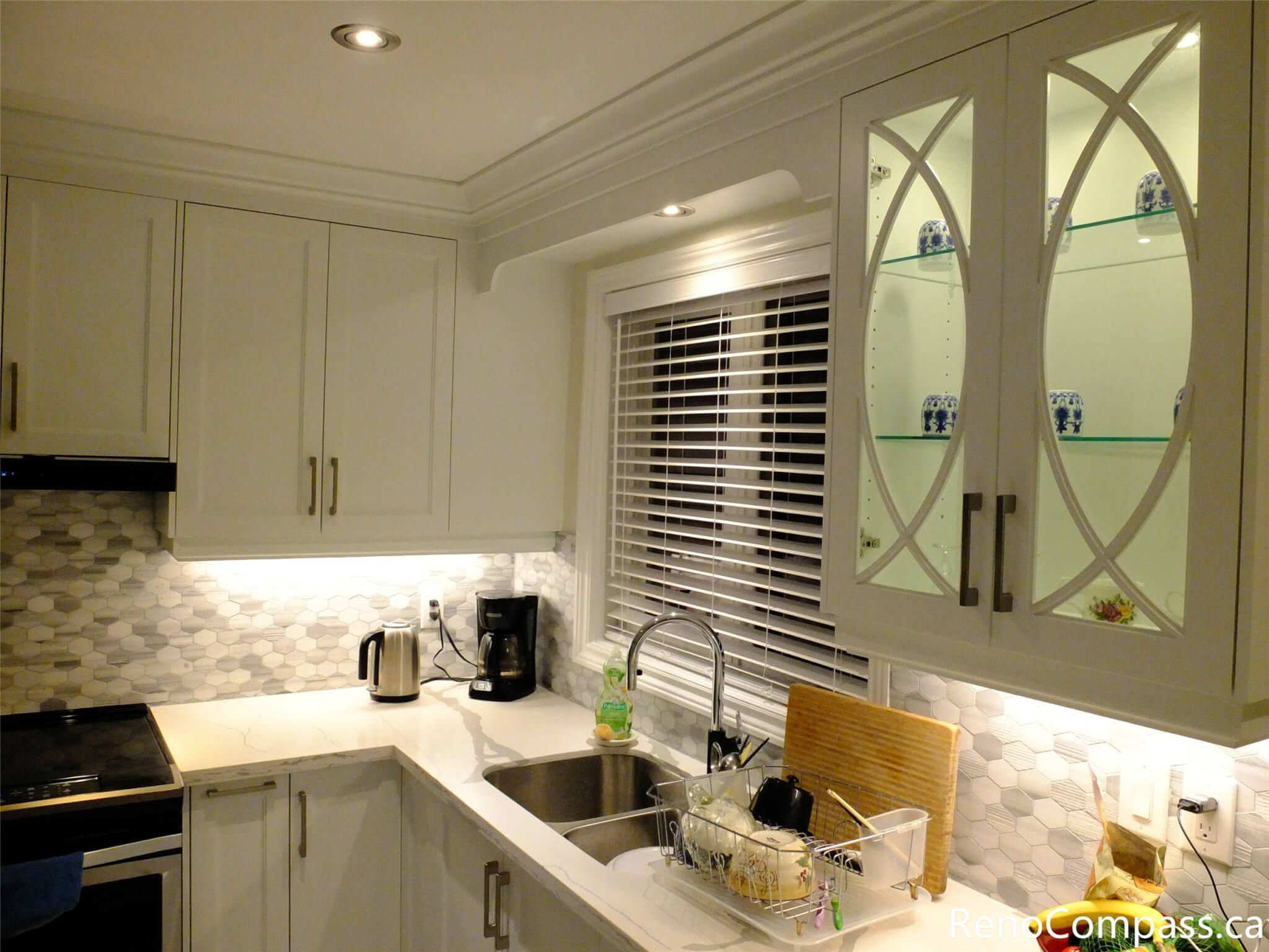 This North York kitchen renovation highlights custom white cabinetry with decorative glass doors, integrated under-cabinet lighting, and a functional L-shaped layout. The quartz countertop, hexagon tile backsplash, and updated sink area create a clean and efficient workspace, completed by a professional Toronto renovation company.