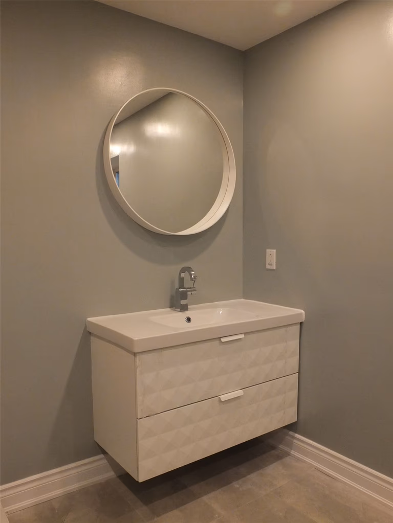 This image highlights a modern bathroom vanity renovation as part of a Mississauga interior renovation project. The design features a floating vanity with integrated drawers, a sleek single-handle faucet, and a round wall-mounted mirror that enhances visual balance. Soft neutral wall tones and minimalist finishes create a clean, contemporary look while maintaining functionality. Completed by a professional Toronto renovation company, this bathroom upgrade improves storage efficiency, space optimization, and overall interior aesthetics for Mississauga homes.