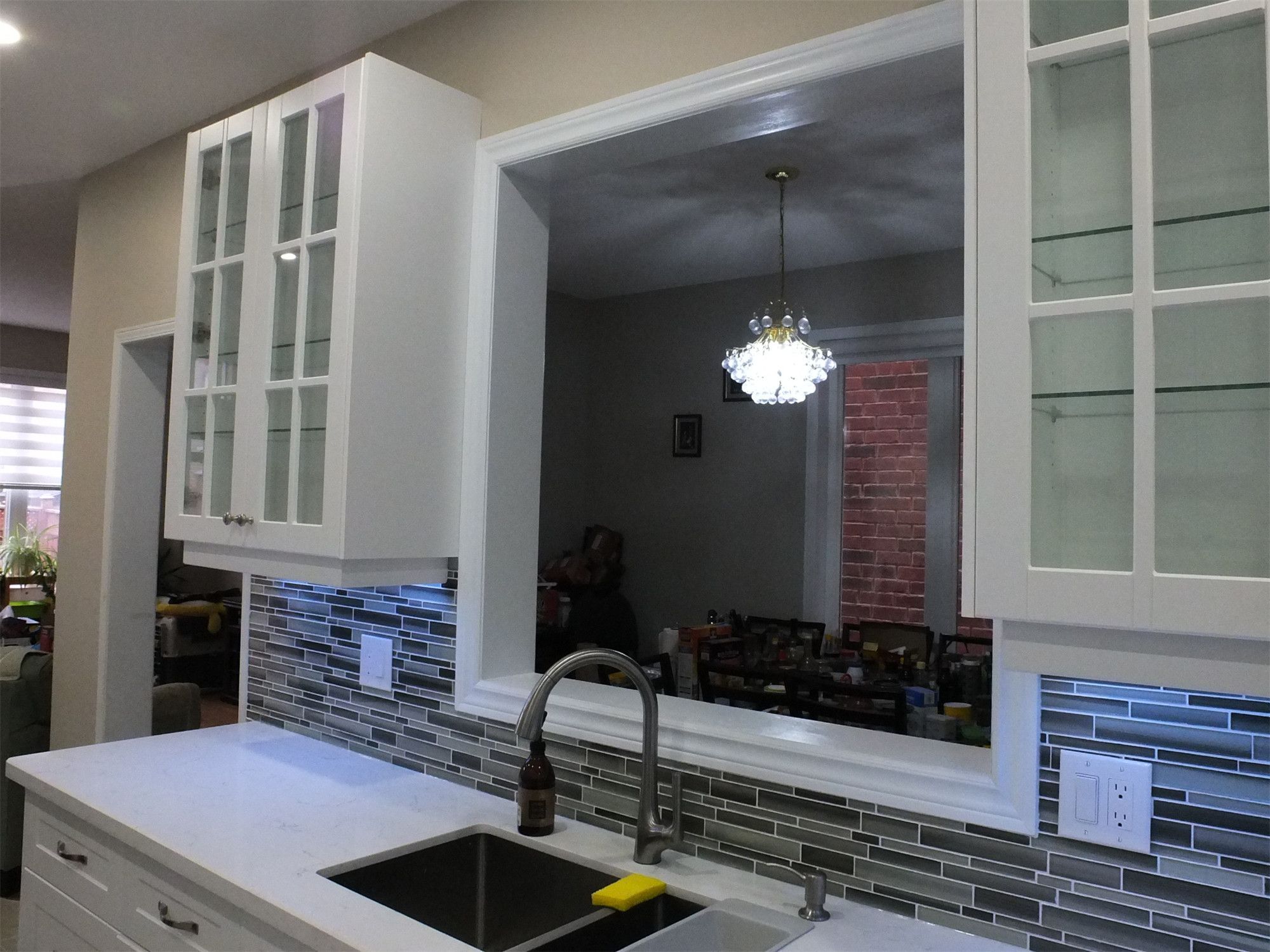 This Vaughan kitchen renovation features elegant white shaker cabinets with glass display doors, quartz countertops, and a stylish mosaic tile backsplash with LED under-cabinet lighting. The pass-through opening above the sink connects the kitchen to the dining area, creating a brighter and more open layout. Completed by a professional Toronto renovation company, the design blends modern functionality with clean, timeless aesthetics.