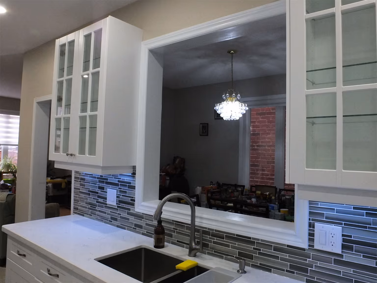 This Vaughan kitchen renovation features elegant white shaker cabinets with glass display doors, quartz countertops, and a stylish mosaic tile backsplash with LED under-cabinet lighting. The pass-through opening above the sink connects the kitchen to the dining area, creating a brighter and more open layout. Completed by a professional Toronto renovation company, the design blends modern functionality with clean, timeless aesthetics.
