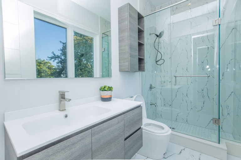 Bright modern bathroom renovation in a 5000 sqft Toronto home featuring a frameless glass shower enclosure, marble-style wall tiles, floating grey vanity with integrated sink, and large mirror reflecting natural light. The clean layout and contemporary finishes create a spacious spa-like atmosphere with functional storage and elegant detailing. Completed by Reno Compass Inc., a professional Toronto renovation company specializing in high-end bathroom and full home interior renovations.