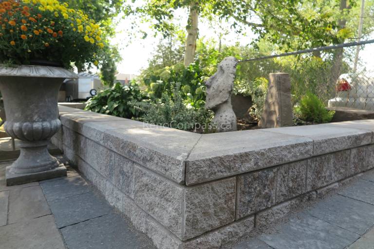 This close-up view highlights the precision craftsmanship of the stone retaining wall and interlocking patio installation in Mississauga. The textured concrete blocks and smooth stone cap provide structural stability while delivering a clean, layered aesthetic. Integrated seamlessly with the surrounding landscaping and planting design, this hardscape feature enhances both durability and visual depth. Professionally completed by Reno Compass Inc., this project reflects expert workmanship in outdoor interlocking and landscape construction.