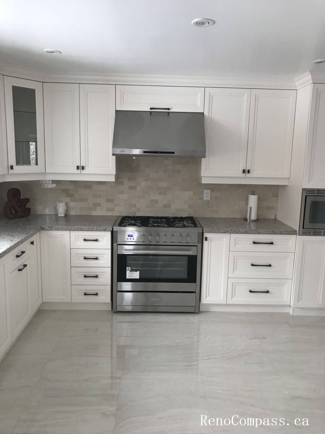 This Markham interior renovation showcases a bright open-concept kitchen featuring custom white cabinetry, stainless steel appliances, and polished porcelain tile flooring. The layout includes a professional-grade gas range with a modern hood fan, built-in microwave, and a full-size stainless steel refrigerator, creating a functional and streamlined cooking space. Neutral mosaic backsplash tiles add subtle texture while maintaining a timeless aesthetic. The clean lines, balanced cabinetry proportions, and durable finishes reflect thoughtful planning and precision installation. This kitchen renovation highlights Reno Compass’ expertise in delivering high-quality interior renovation services across Markham and the Greater Toronto Area.