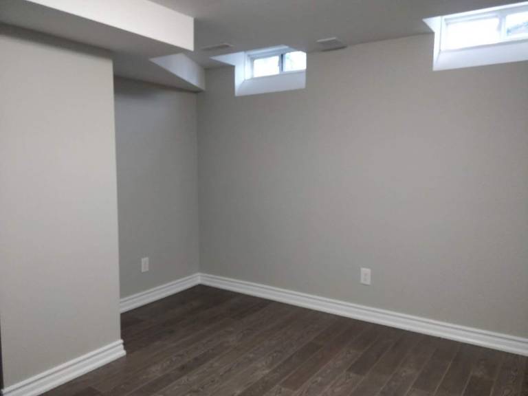 This image features a finished basement room from a Scarborough renovation project, showcasing newly installed dark hardwood flooring, fresh neutral wall paint, and upgraded baseboards for a clean, modern appearance. The space includes a standard entry door and mirrored closet doors, creating a functional layout suitable for a bedroom, home office, or guest suite. The crisp trim detailing and cohesive color palette enhance the overall brightness and comfort of the basement. Completed by Reno Compass Inc., this renovation highlights quality finishes and thoughtful interior upgrades.
