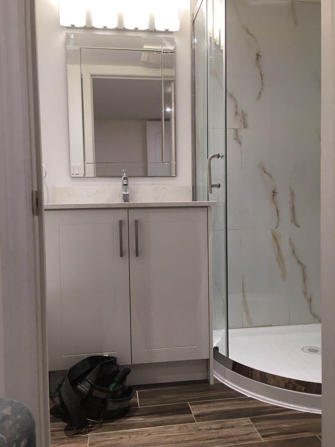 This Markham basement renovation features a contemporary basement bathroom with a sleek white vanity, modern mirror lighting, and a curved glass shower enclosure with marble-look wall panels. The warm-toned flooring and clean finishes create a bright, elegant, and functional bathroom space tailored for Markham homeowners.
